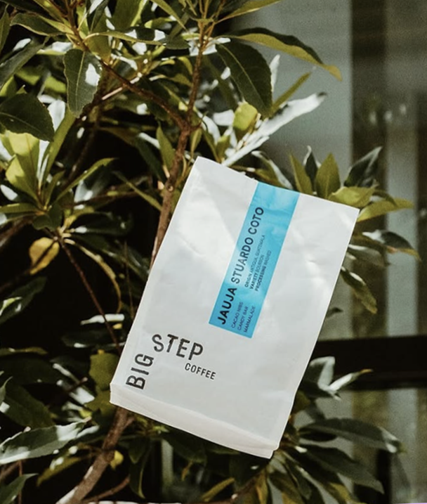 A coffee bag is being thrown into the air before a green leafy shrub with sunlight.