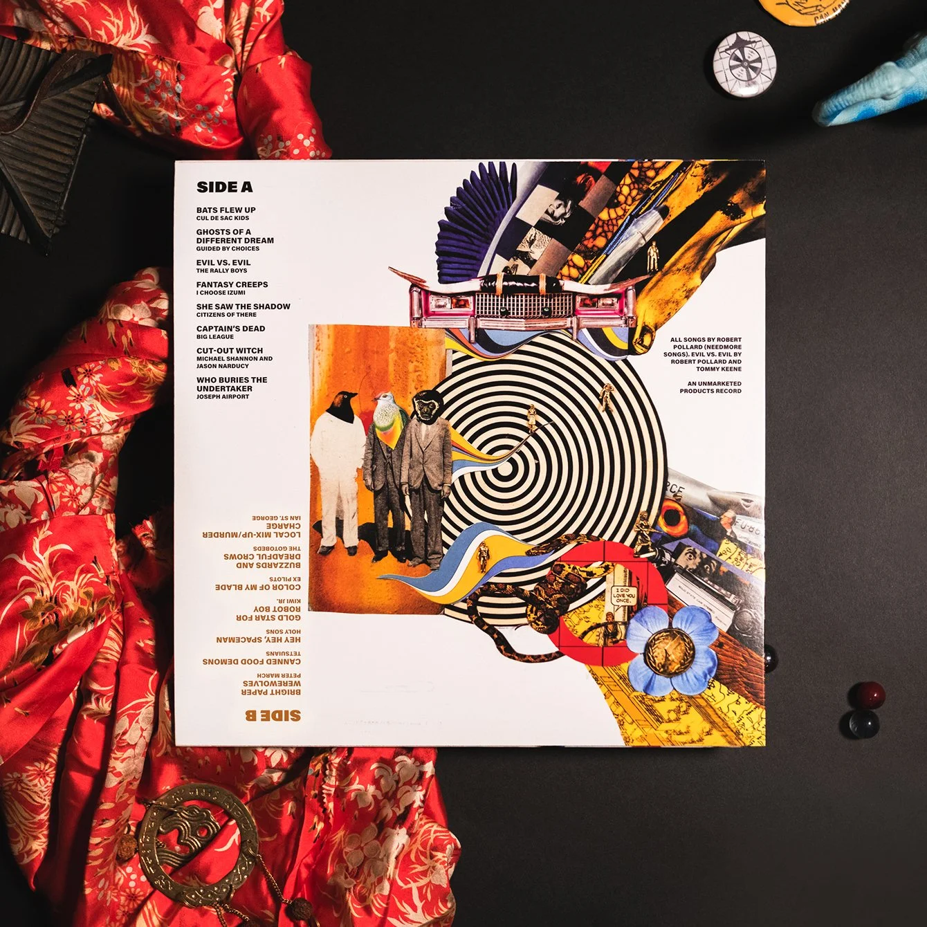 The back cover of a vinyl record album, featuring a colorful, surreal collage with a spiral design, animals with human bodies, and various objects, laid on a black surface next to red and blue fabric and small decorative items.
