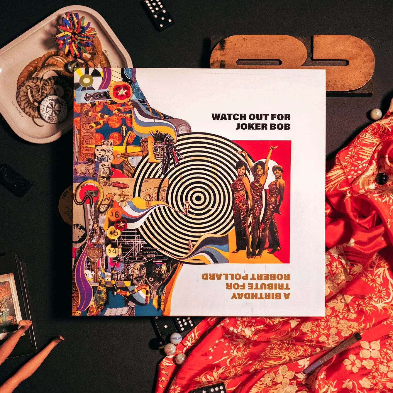 A vinyl record album cover titled 'Watch Out for Joker Bob,' featuring women in sparkly outfits, a black and white hypnotic spiral, and various collage elements including cartoon characters and vintage images, on a dark surface with scattered objects like dominoes, a toy, and a red floral fabric.