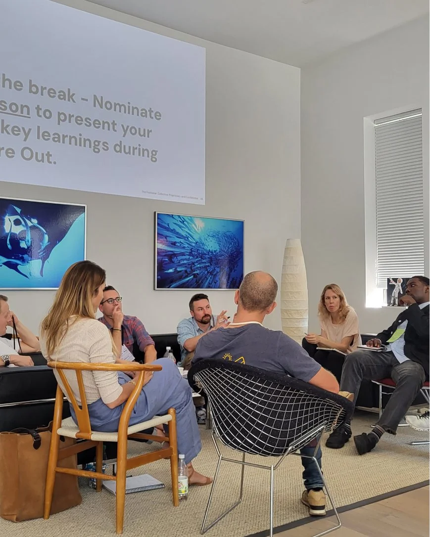 A group of people sitting in a circle in a room with white walls and multiple screens, engaged in a discussion or meeting.