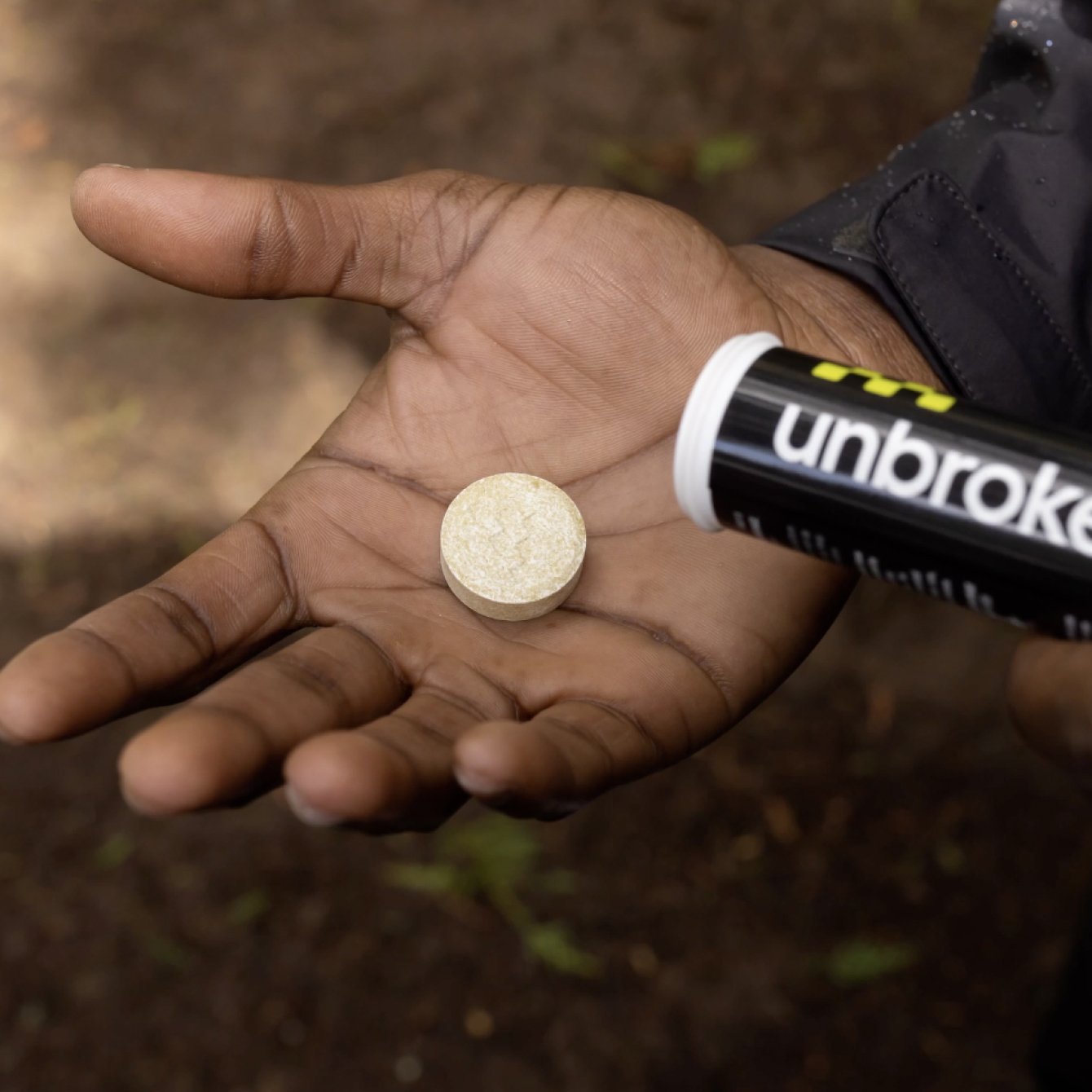 A hand holding a round beige dissolvable tablet with a black and white tube labeled 'unbroken' nearby, in a natural outdoor setting.