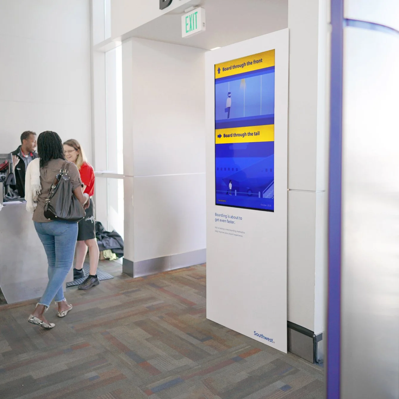 Airport gate area with a group of three people at the check-in counter, a digital boarding sign, and an emergency exit sign overhead.