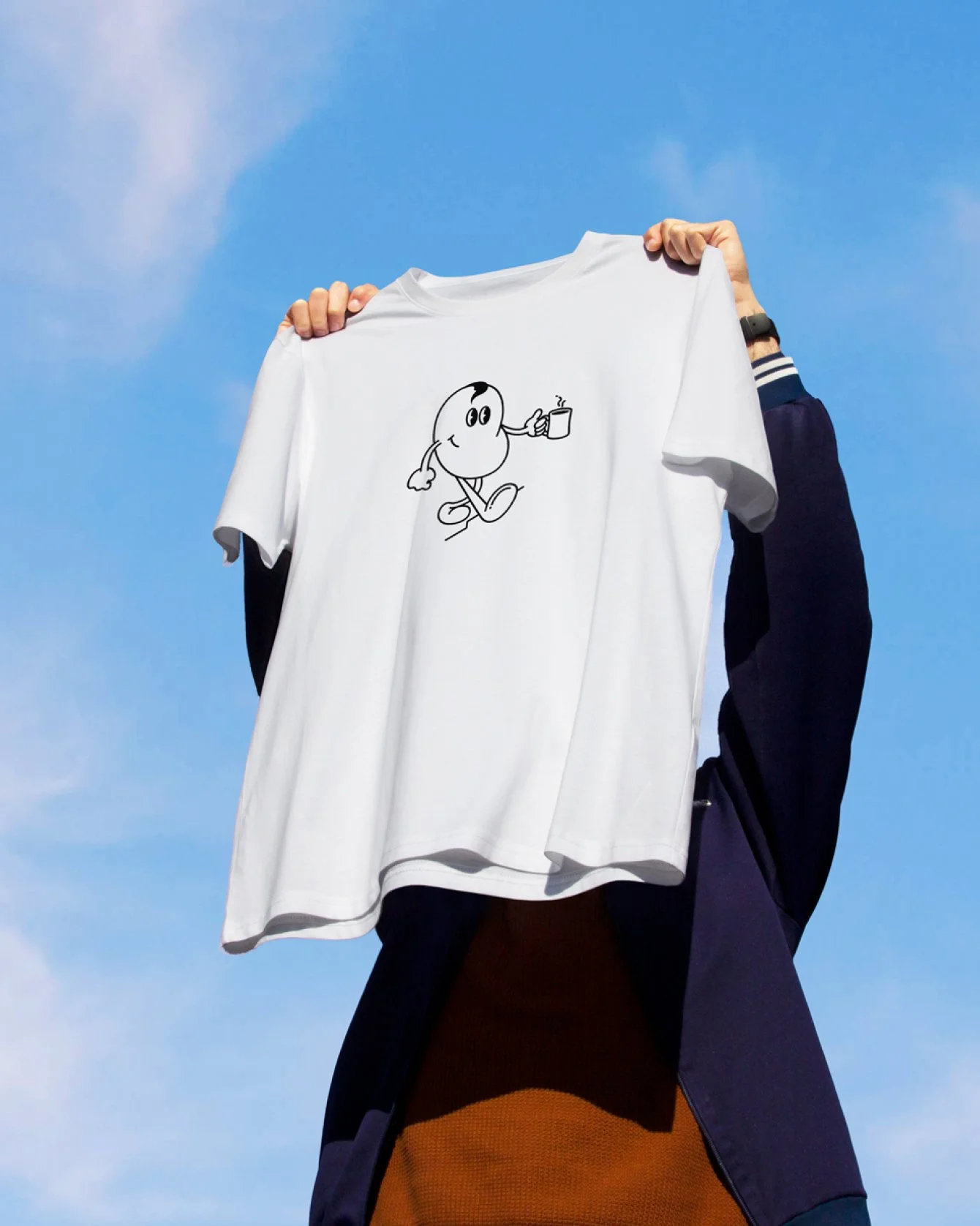 Person holding up a white T-shirt with a black cartoon character drinking from a mug, against a blue sky background.