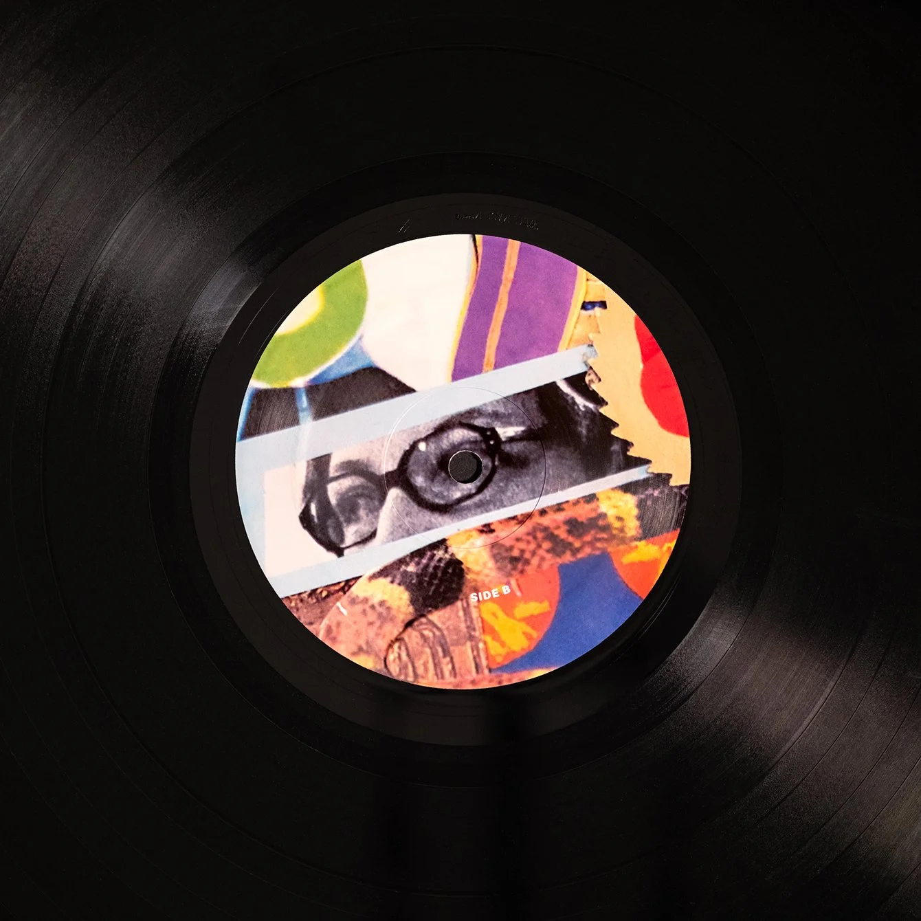 Close-up view of a vinyl record with a colorful collage-style label featuring a woman's face, glasses, and various abstract artwork.