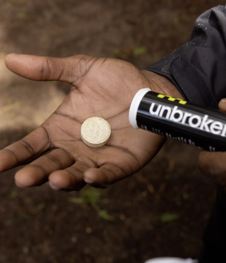 A person holding a large, round, dissolvable tablet with their left hand, while using an unbroken branded tube with a yellow and black design in their right hand. The background is outdoors with a blurred, earthy ground.