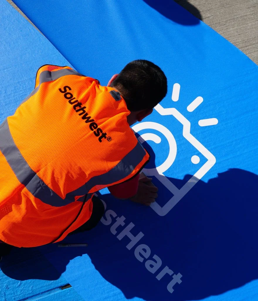A worker in an orange vest with 'Southwest' on the back is kneeling on a large blue surface and appears to be doing some work on a graphic or logo that includes a stylized heart, a camera, and the word 'Heart'.