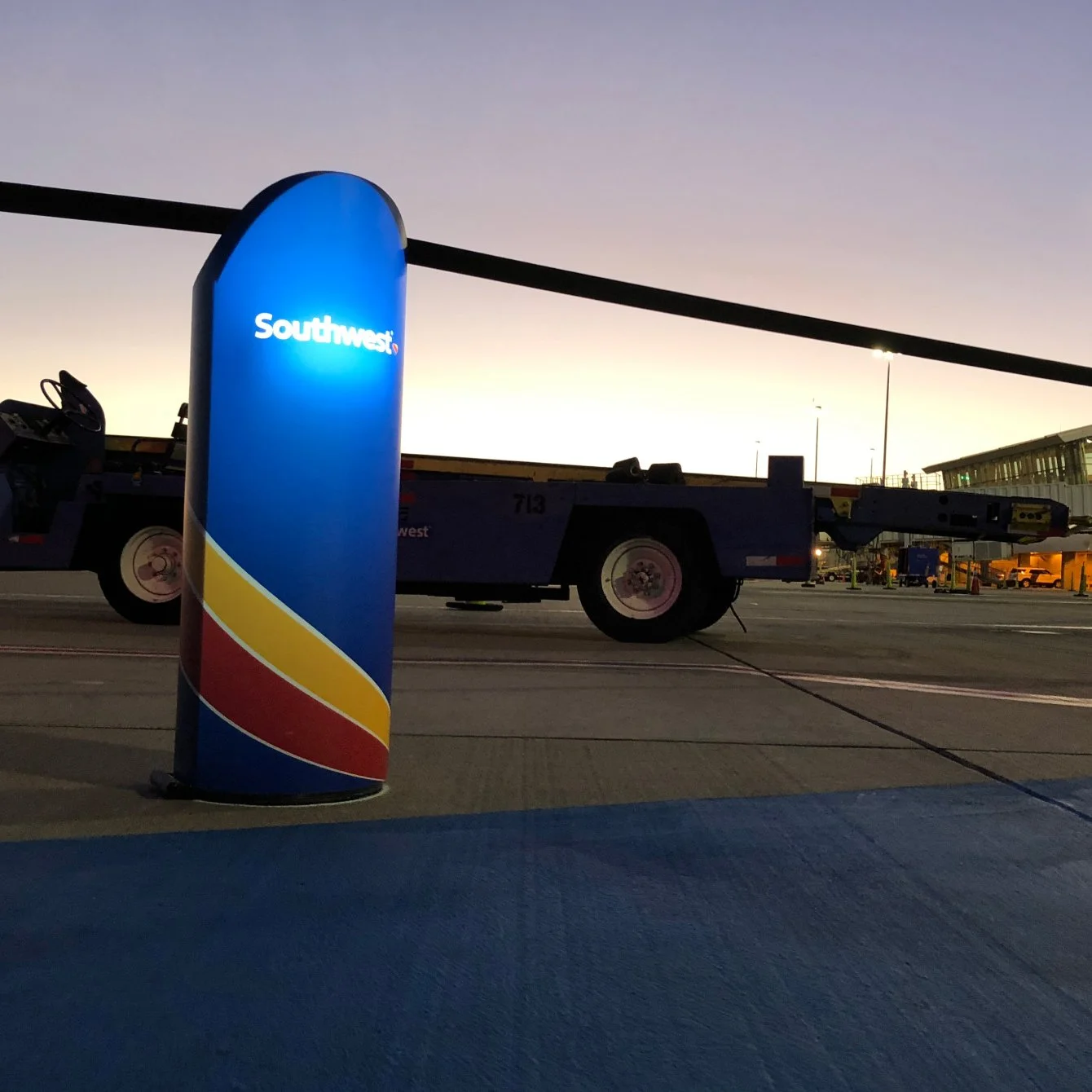 At an airport gate during dusk, a blue Southwest Airlines stanchion with red, yellow, and dark blue swoosh design is illuminated. A jet bridge and ground support equipment are visible in the background.