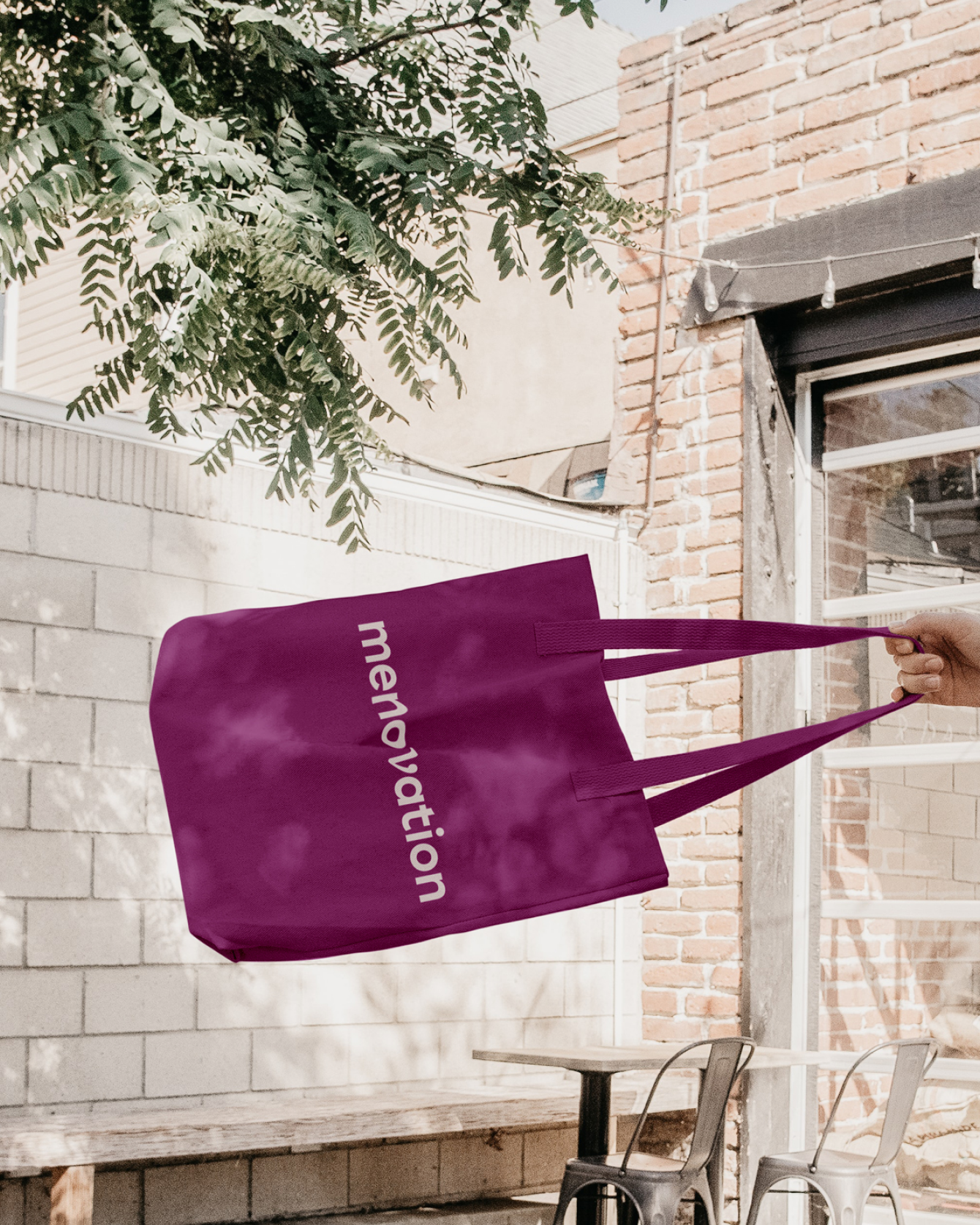 Purple tote bag with the word 'menlovenation' being held by a person's hand outdoors, with a brick building, a concrete wall, a table, and chairs in the background.