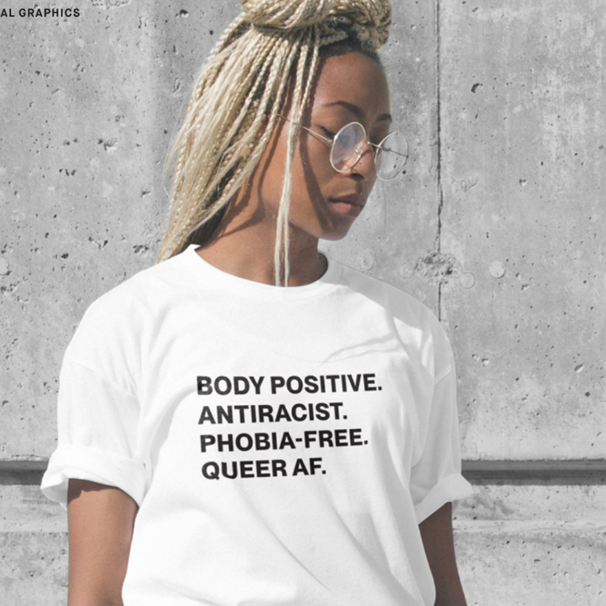 A woman with long blonde dreadlocks and glasses wearing a white T-shirt with bold black text that reads: "BODY POSITIVE. ANTIRACIST. PHOBIA-FREE. QUEER AF." standing against a concrete wall.
