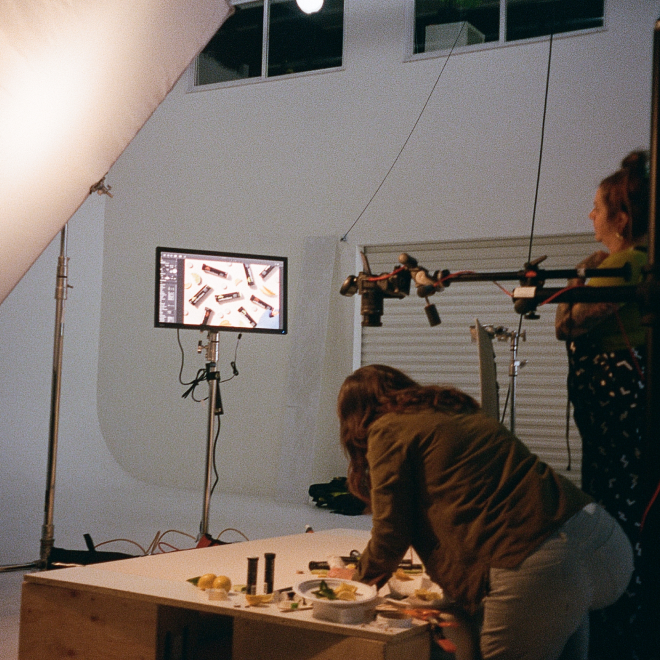 Photo of a food photo shoot setup indoors with two women preparing dishes on a table, studio lighting, and a monitor displaying the styled dishes.