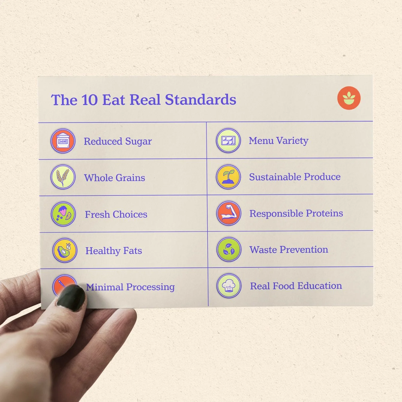 A hand with black painted fingernails holding a list titled 'The 10 Eat Real Standards' featuring icons and points such as Reduced Sugar, Whole Grains, Fresh Choices, Healthy Fats, Minimal Processing, Menu Variety, Sustainable Produce, Responsible Proteins, Waste Prevention, and Real Food Education.