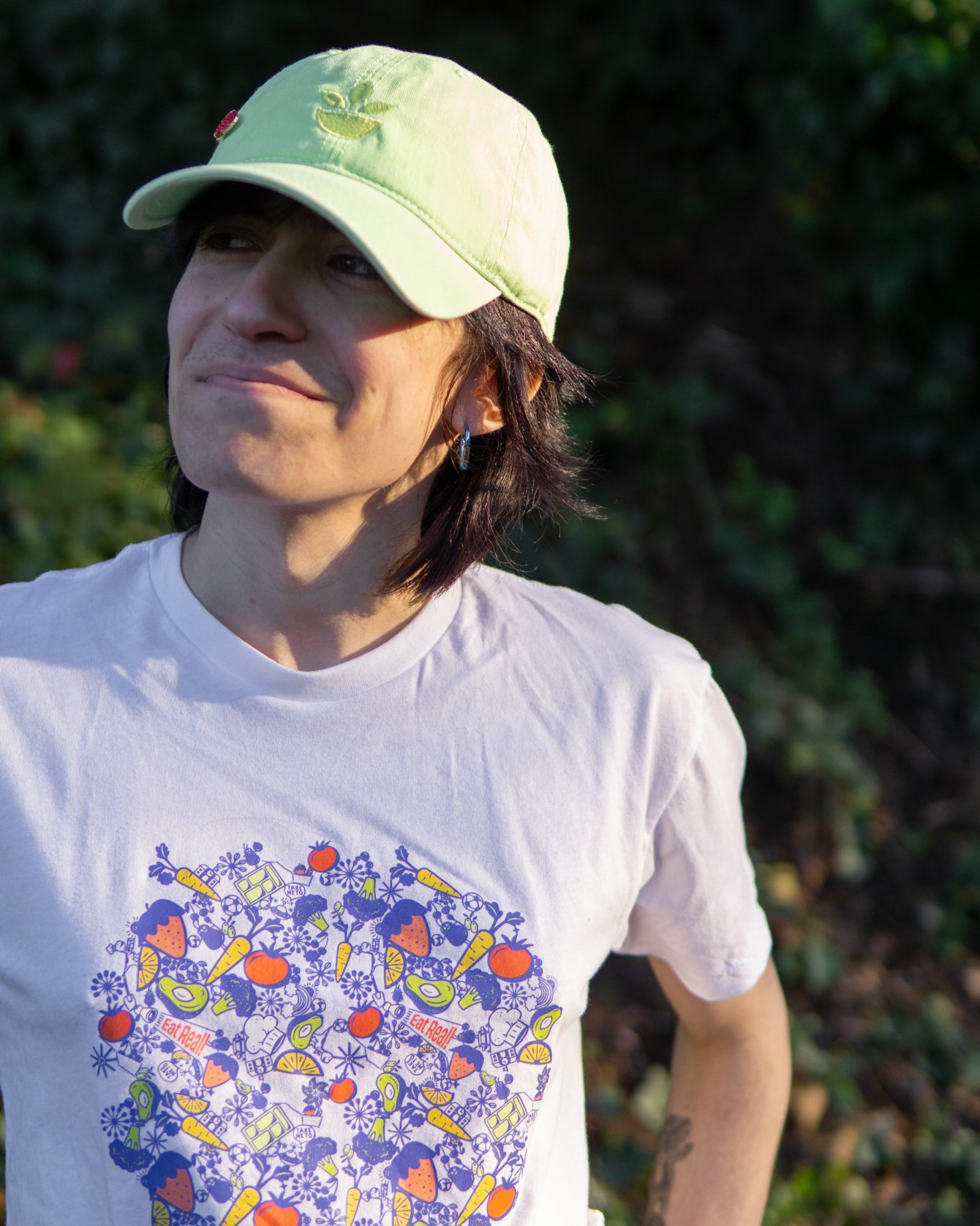 A non-binary person wearing a light green baseball cap and a white T-shirt with colorful fruit and vegetable illustrations and the phrase "Eat Real!" standing outdoors with greenery in the background.