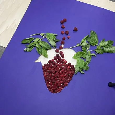 A craft project on a purple background using raspberries and green leaves to create the shape of a strawberry.