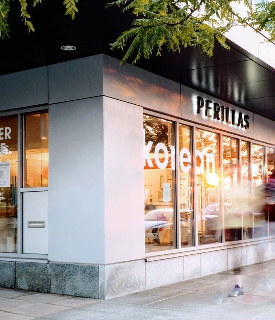 Storefront with large window reflecting sunlight and trees, sign reading 'PERILLAS' above the window, and a neon sign on the window that says 'Kore' and 'kitchen.' A person in motion appears as a blur in front of the window.