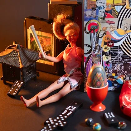 A Barbie doll with blonde hair and a pink dress sitting among various colorful objects and toys, including a remote control, a small house, a decorative egg, and a wall collage with photos and drawings.