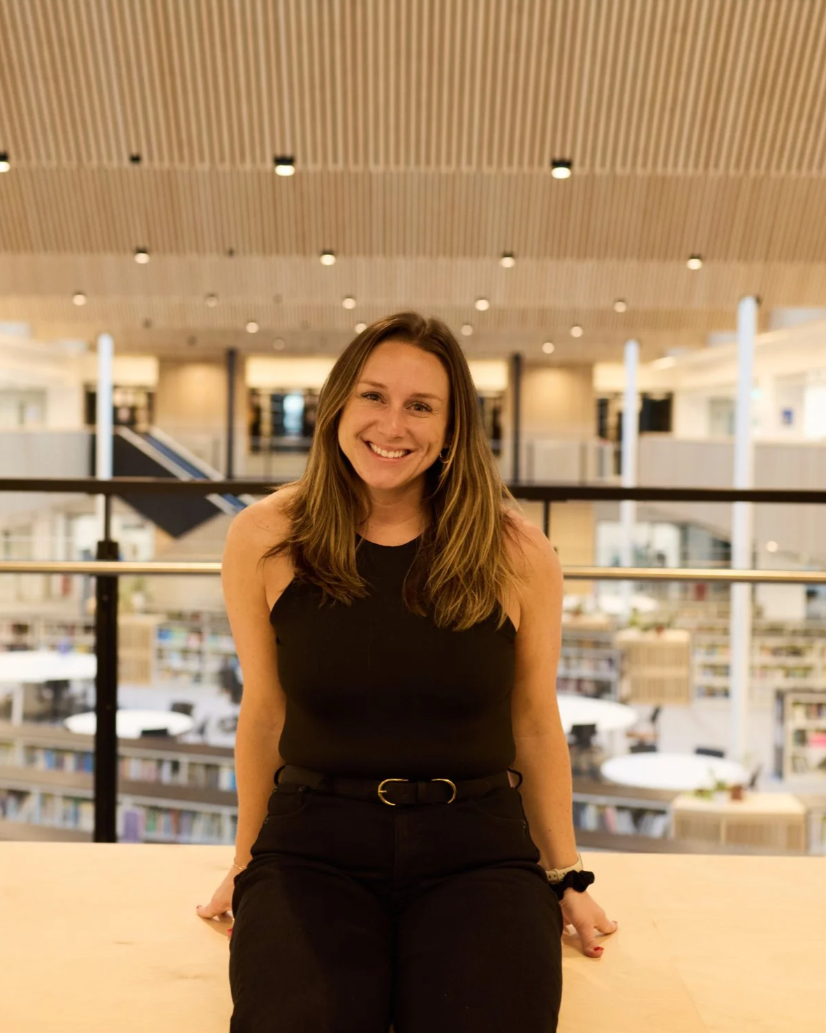Kelly Benson, smiling, wearing a black sleeveless top and black pants, sitting at a table in a library.