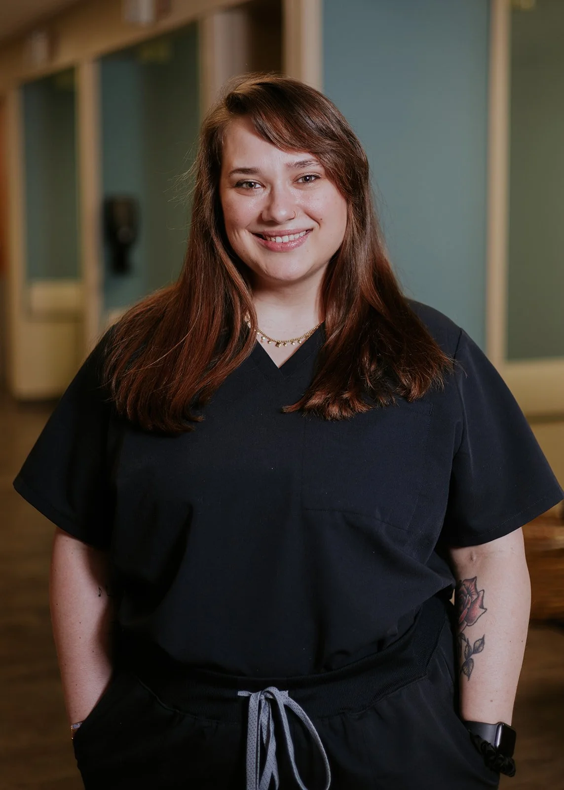 Smiling woman with curly hair standing indoors wearing a black shirt, showing tattoos on her arms.