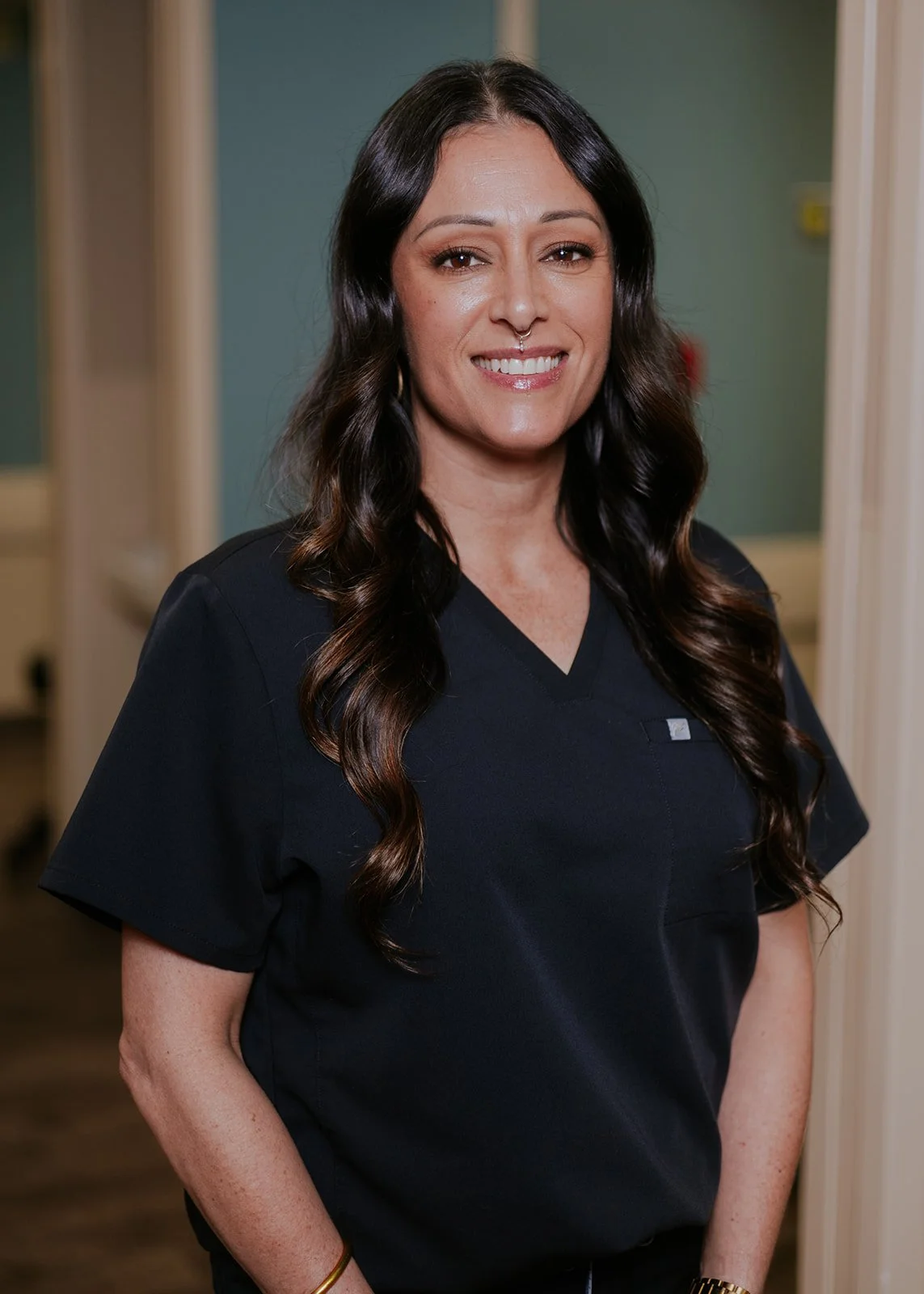 Smiling woman with long dark hair and hoop earrings, wearing a black shirt and smart watch, standing in an indoor setting.