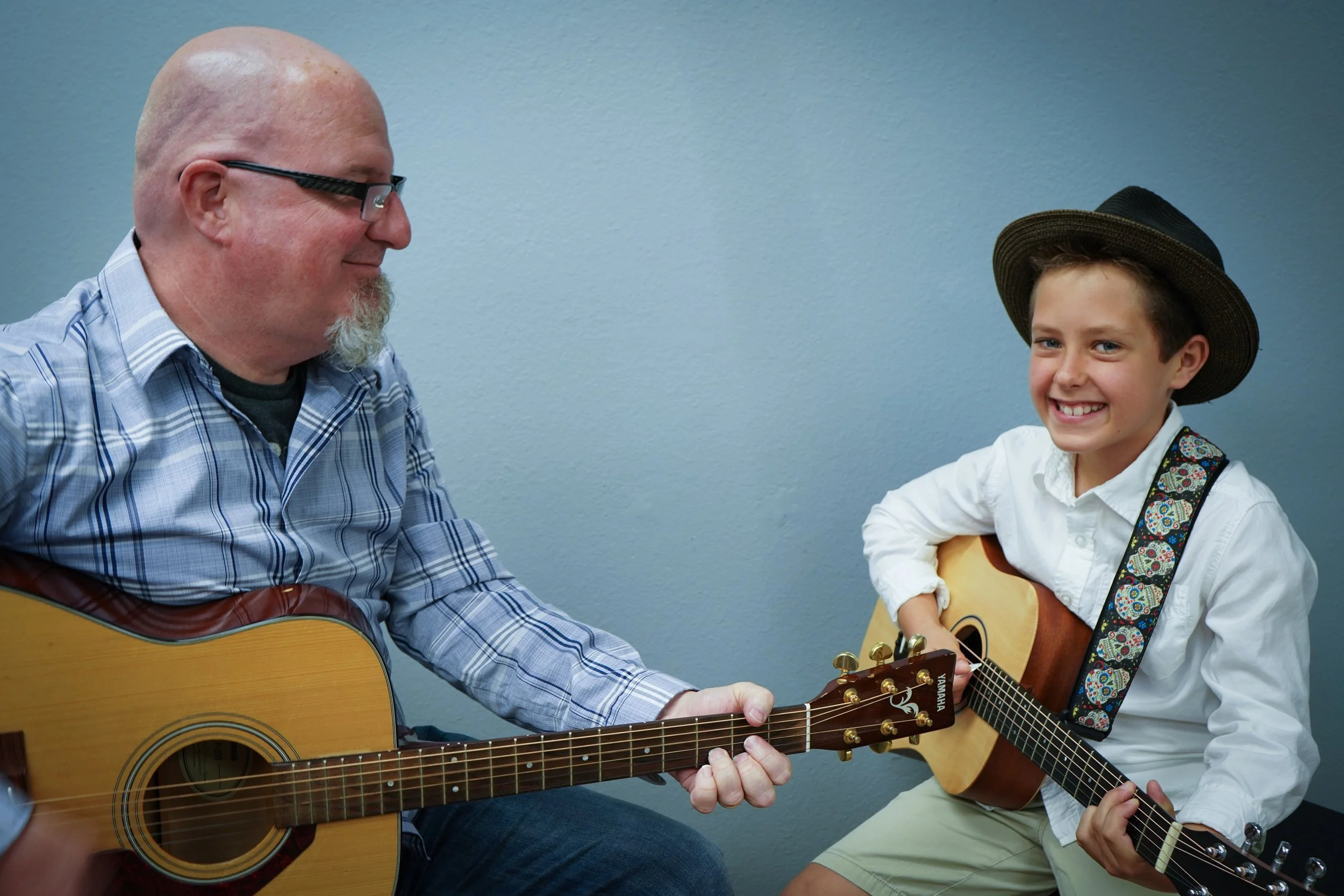 A father and son playing guitars, both smiling. The father is wearing glasses and a plaid shirt, while the son is in a white shirt, khaki shorts, and a wide-brimmed hat.