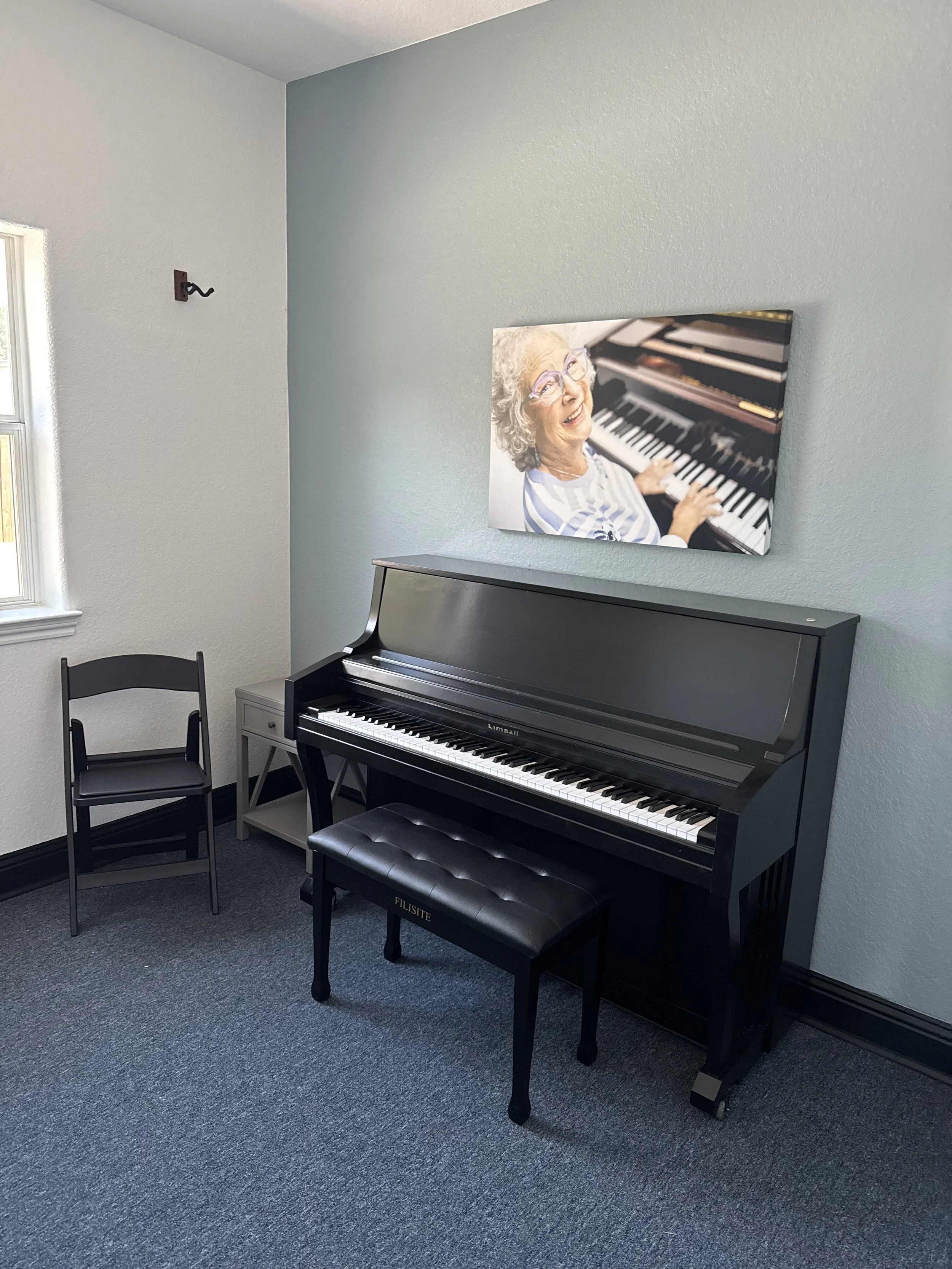 A black upright piano with a matching bench in a room with gray walls and a gray carpet. A chair is next to the piano, and a framed photo of an elderly woman playing the piano is hanging on the wall above it.