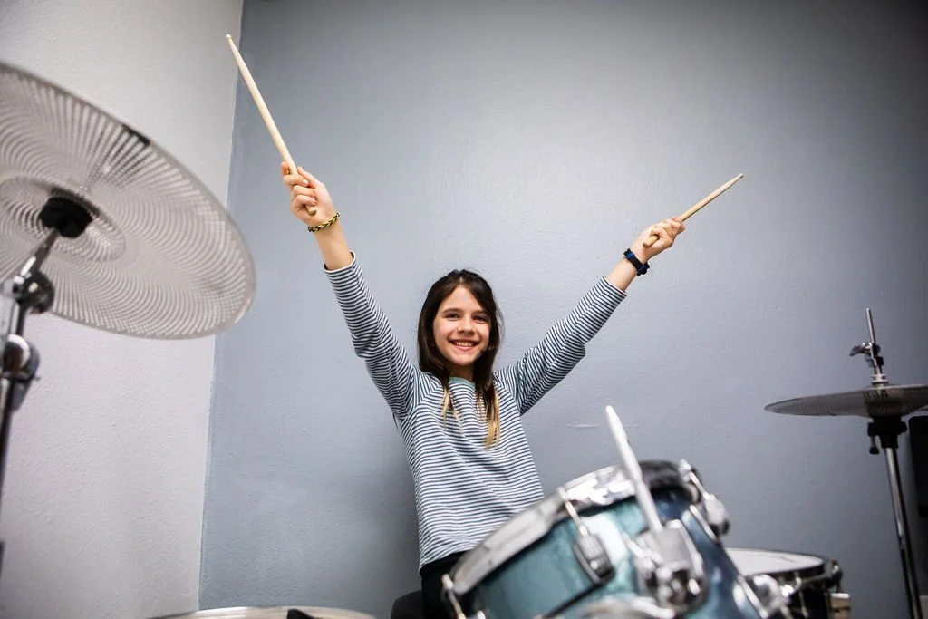 A young girl in a striped shirt smiling and raising drumsticks in front of a drum set in a music room.
