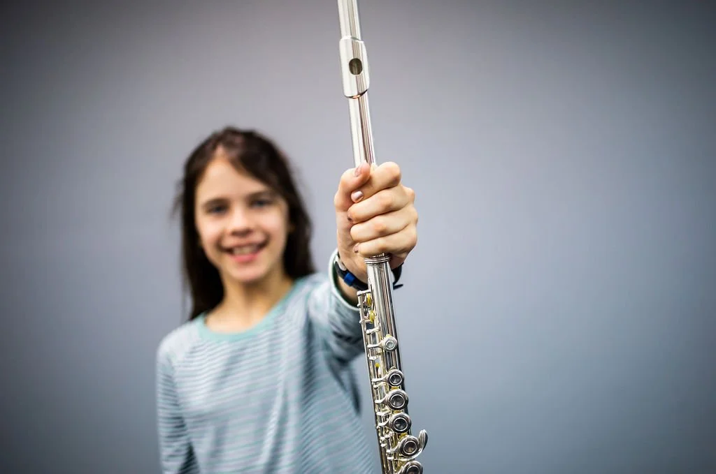 Young girl smiling and holding a silver flute toward the camera against a plain gray background.