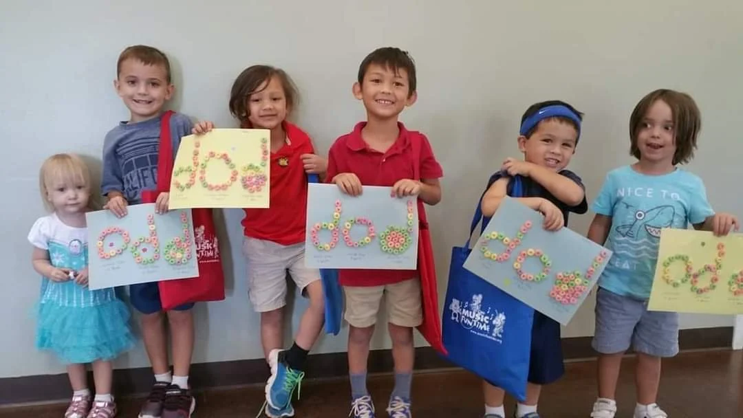 Seven children standing in a row holding colorful handmade signs with the word "Joy" written in flowers.