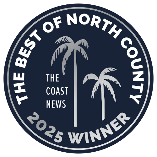 A circular badge with white and gray text on a dark background, featuring two palm trees, with text that reads 'The Best of North County, The Coast News, 2025 Winner.'