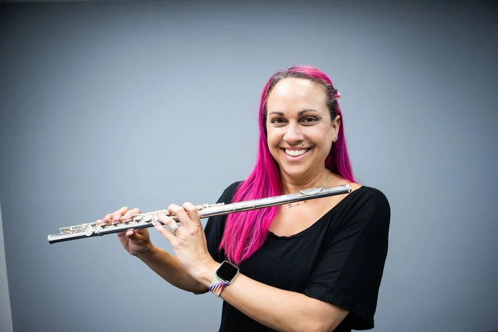 A woman with pink hair holding a silver flute, smiling at the camera against a plain gray background.
