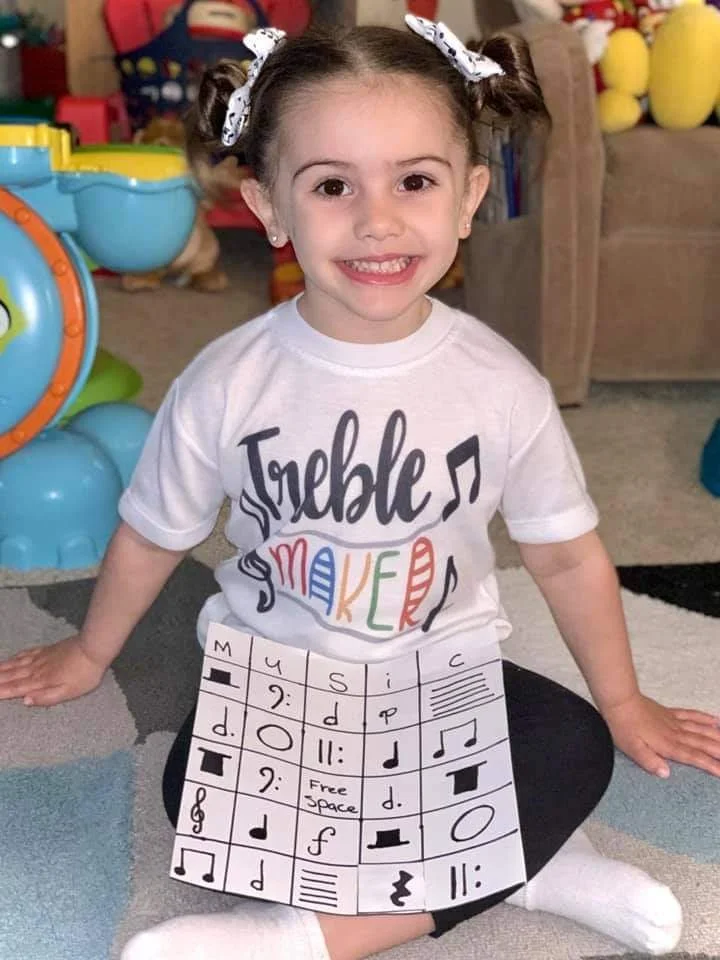 A young girl with brown hair styled in pigtails with white bows, smiling and sitting cross-legged on the floor. She is wearing a white T-shirt with the words 'Treble Maker' and colorful musical notes printed on it. In front of her, there is a sheet o