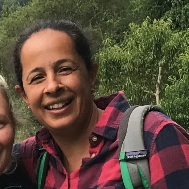 Amrita from California smiling outdoors, wearing a red and black plaid shirt and a gray backpack, with trees in the background.