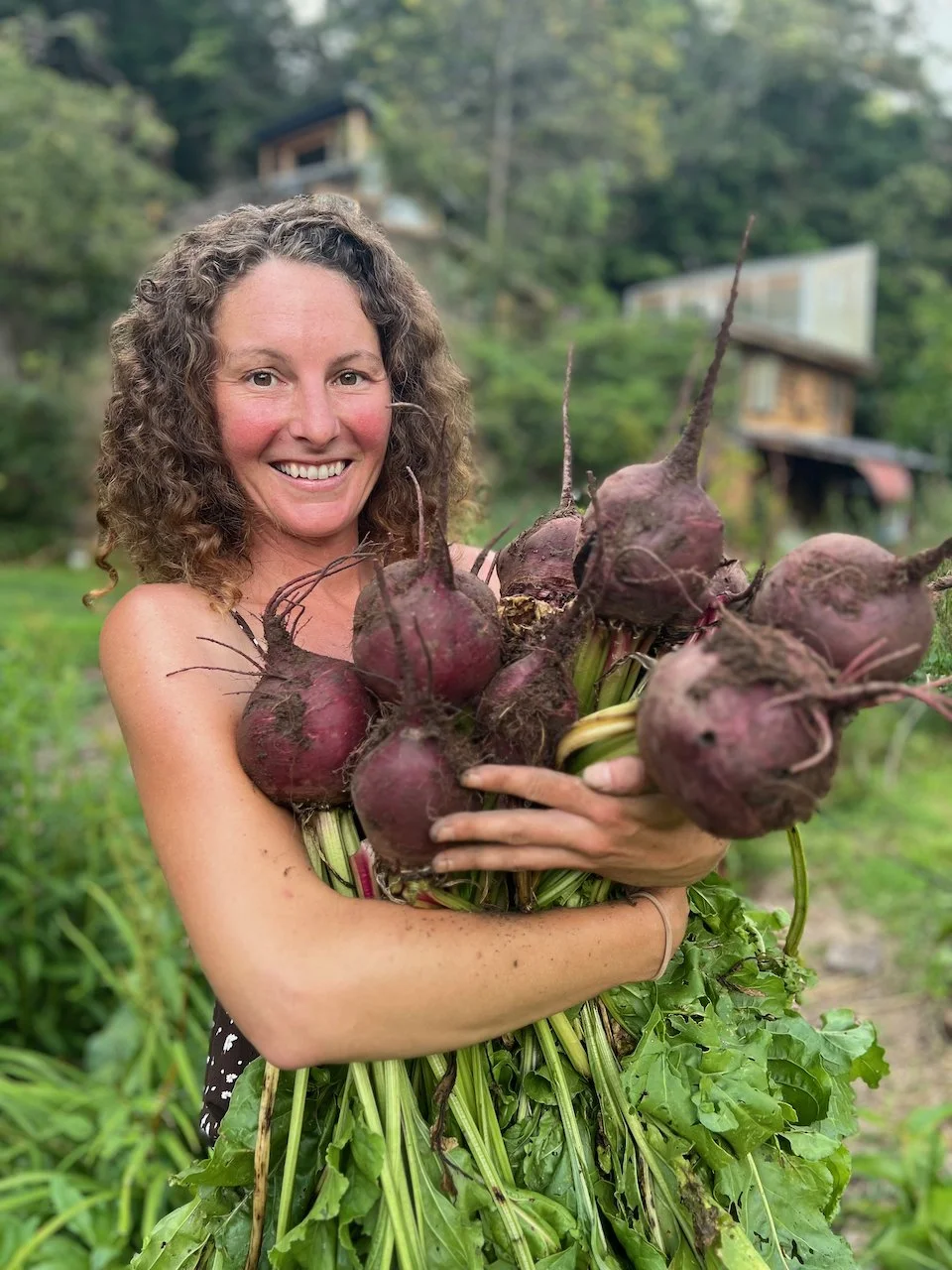 A smiling white woman with curly hair holding a bunch of freshly harvested beets outdoors in a garden.