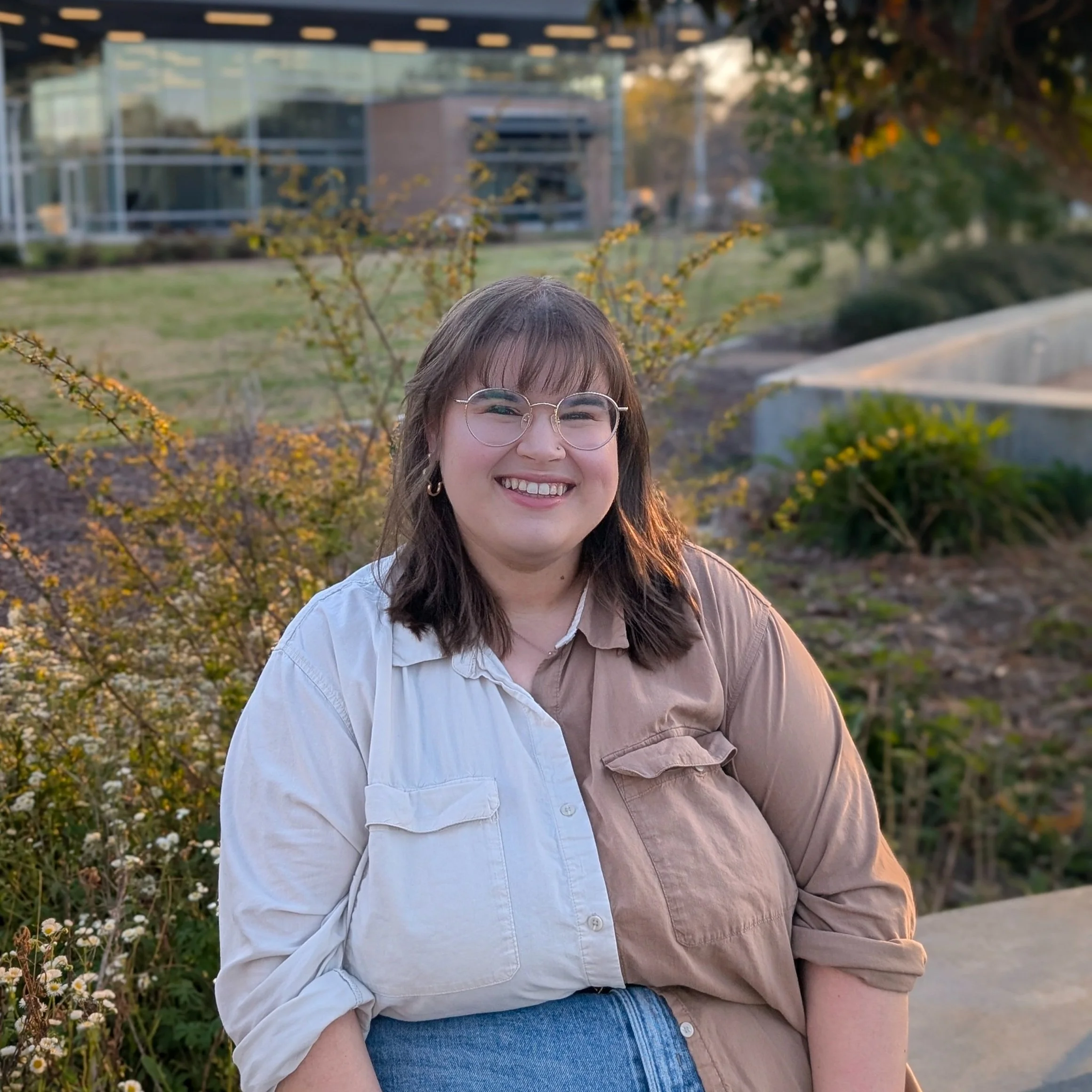 A smiling woman with glasses and brown hair, wearing a button-up shirt, posing outdoors in front of bushes and a building
