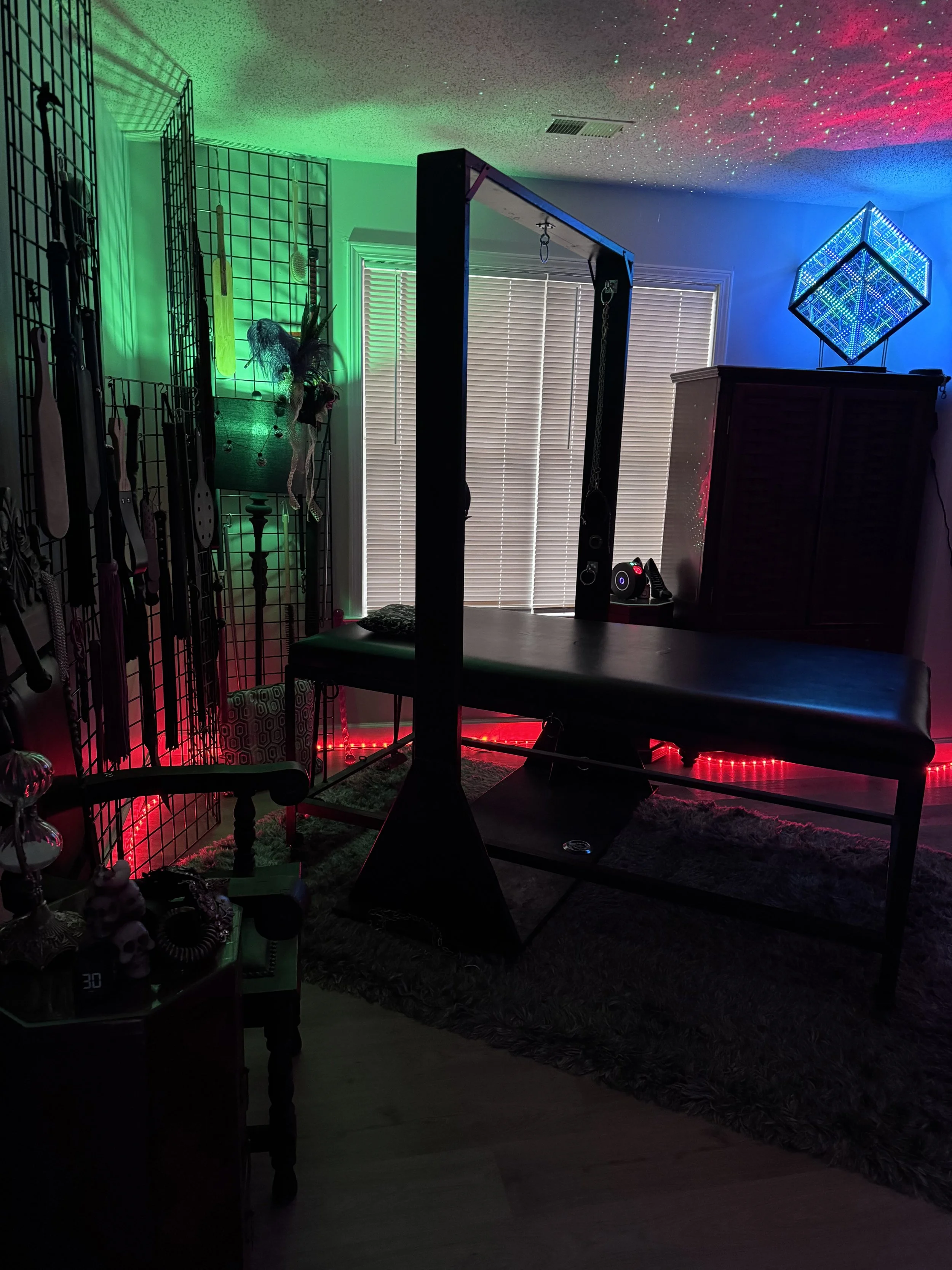 A room with a massage table, colorful LED lights, and fitness equipment, including a squat rack and resistance bands, illuminated by red, green, and blue lighting, and decorated with a glowing geometric light fixture.