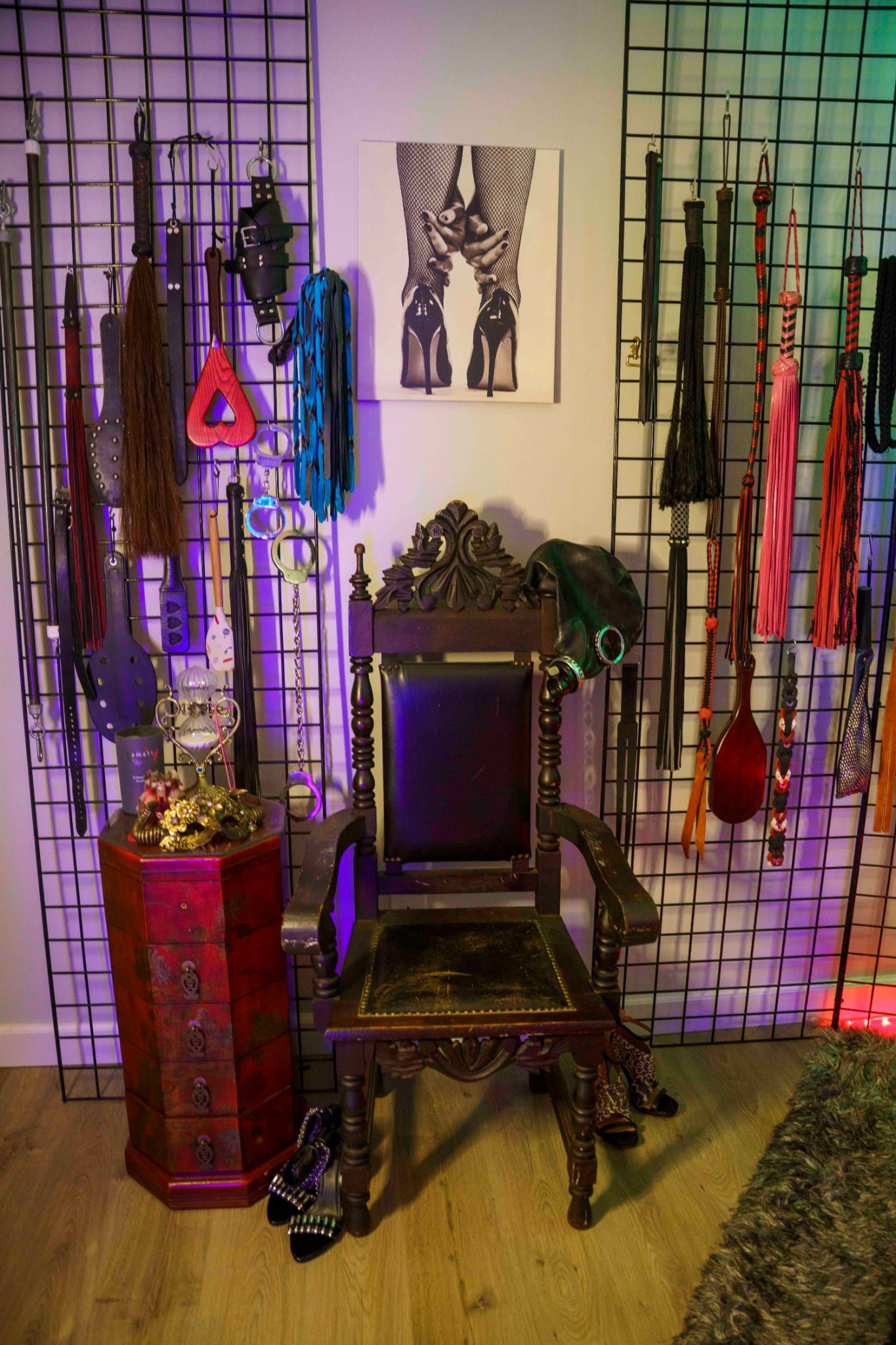 A decorative room wall featuring black grid panels filled with various flogging and BDSM accessories such as whips, paddles, and collars. An ornate wooden chair is centered in front of the wall, with a pair of high-heeled shoes as a decoration on the