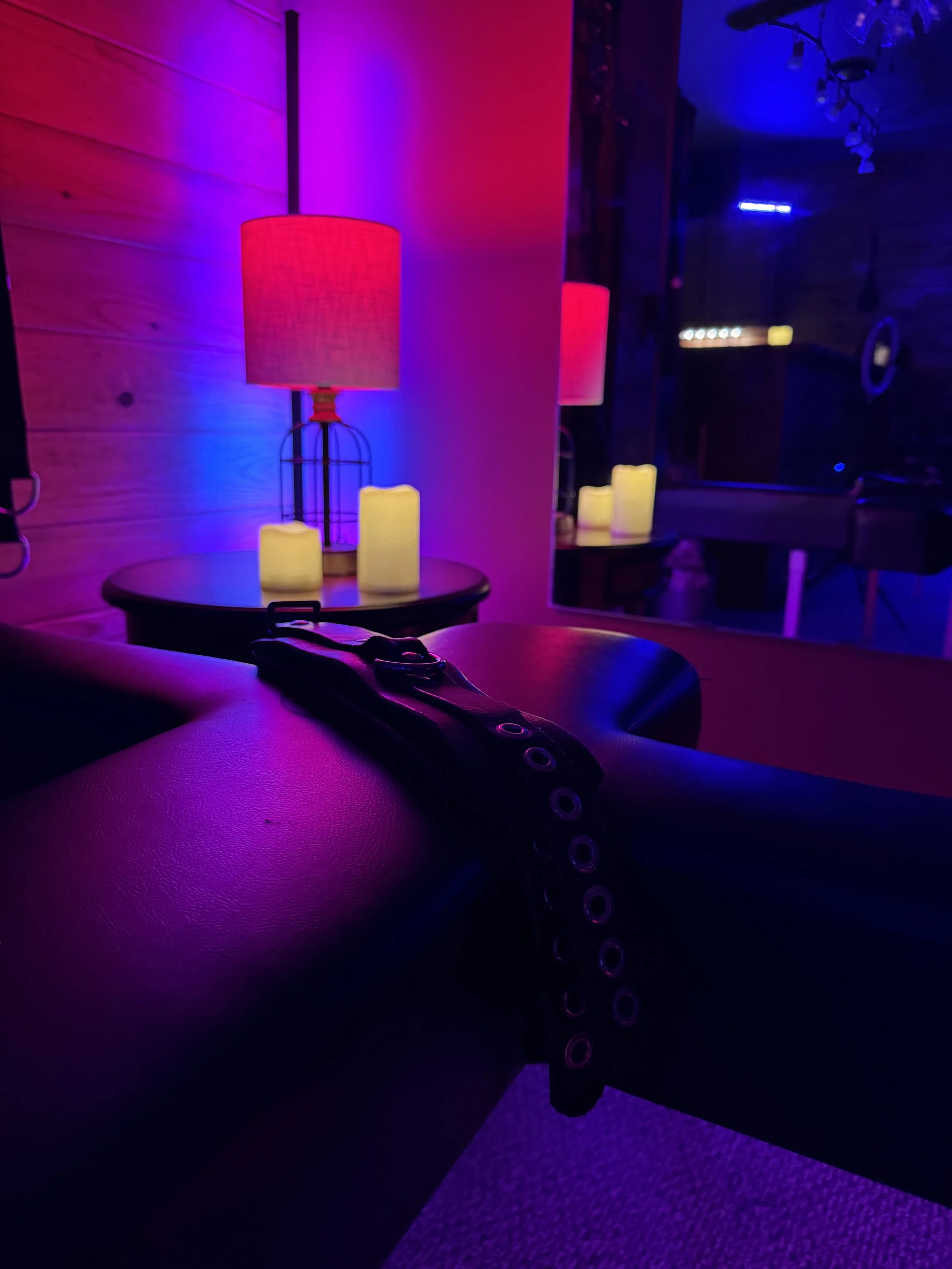 A dark room illuminated by purple, pink, and yellow lighting. A table with a pink lampshade, three yellow candle-like lights, and a mirror reflecting the scene. A belt with metal eyelets is draped over a person's leg in the foreground.
