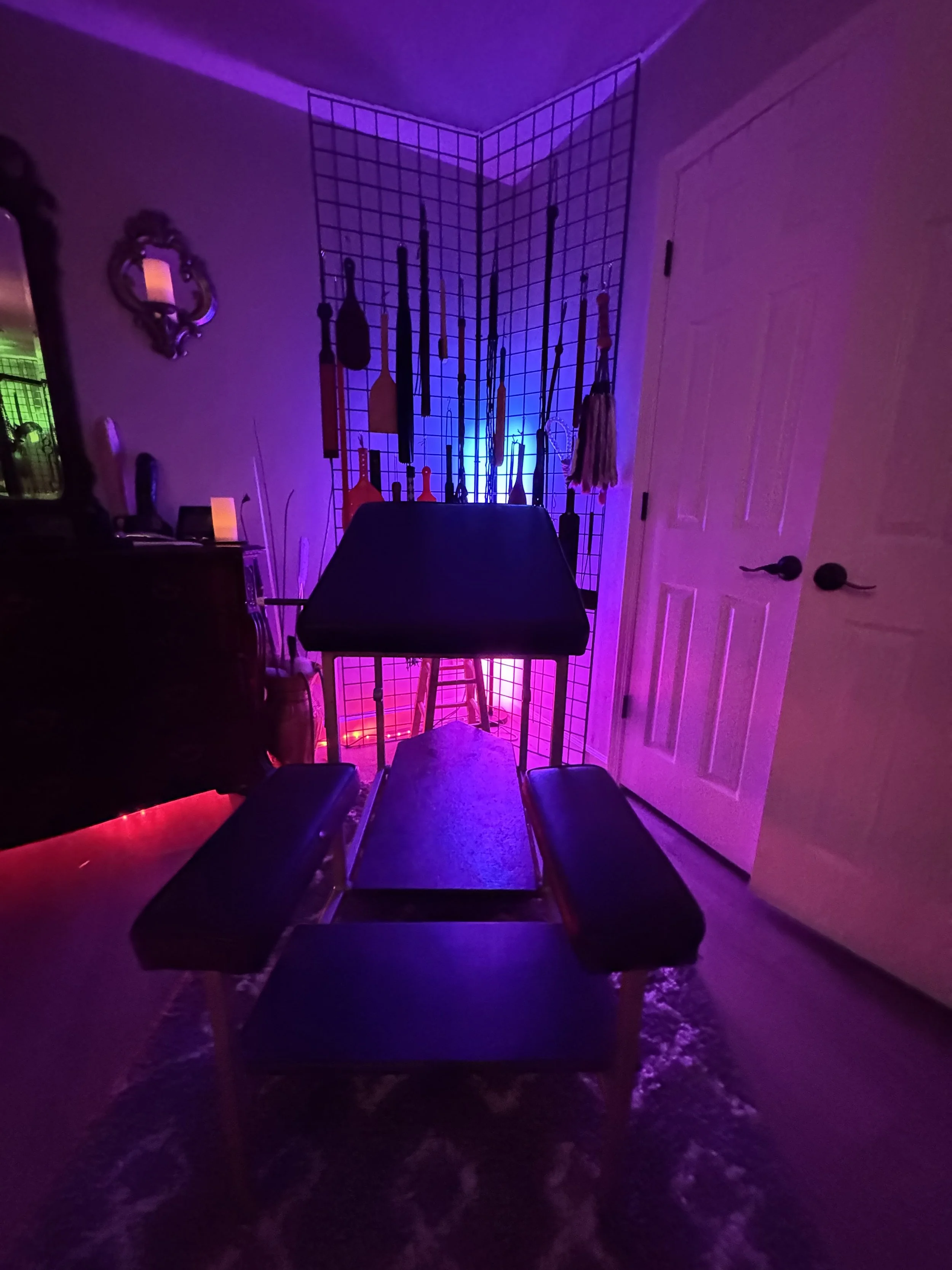 Room with a grid wall display holding various tools, illuminated by colorful purple and pink lights, with a bench in the foreground.