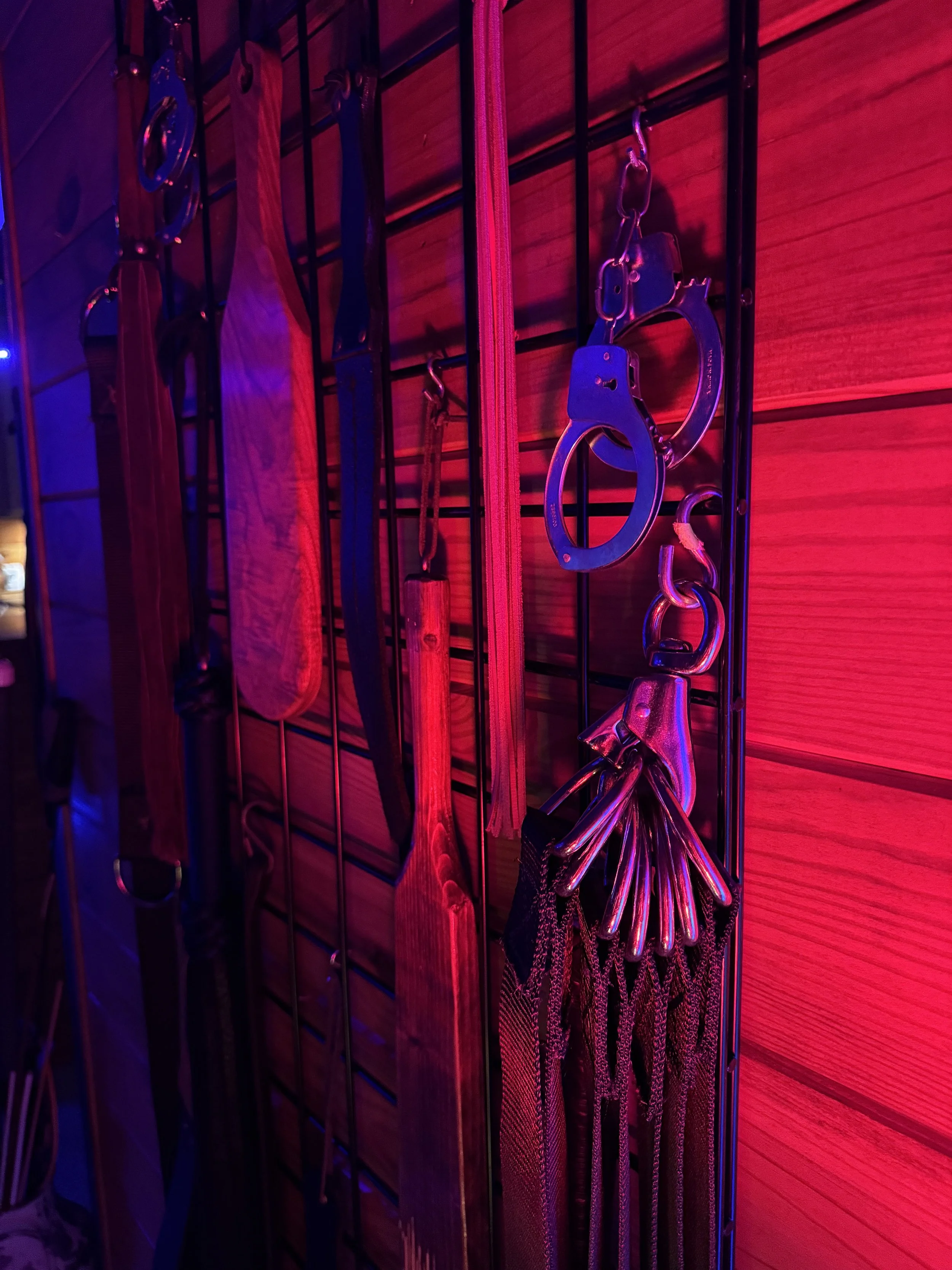 A black metal grid wall with restraints, a leather paddle, and various metal keys hanging, illuminated by red and purple lighting.