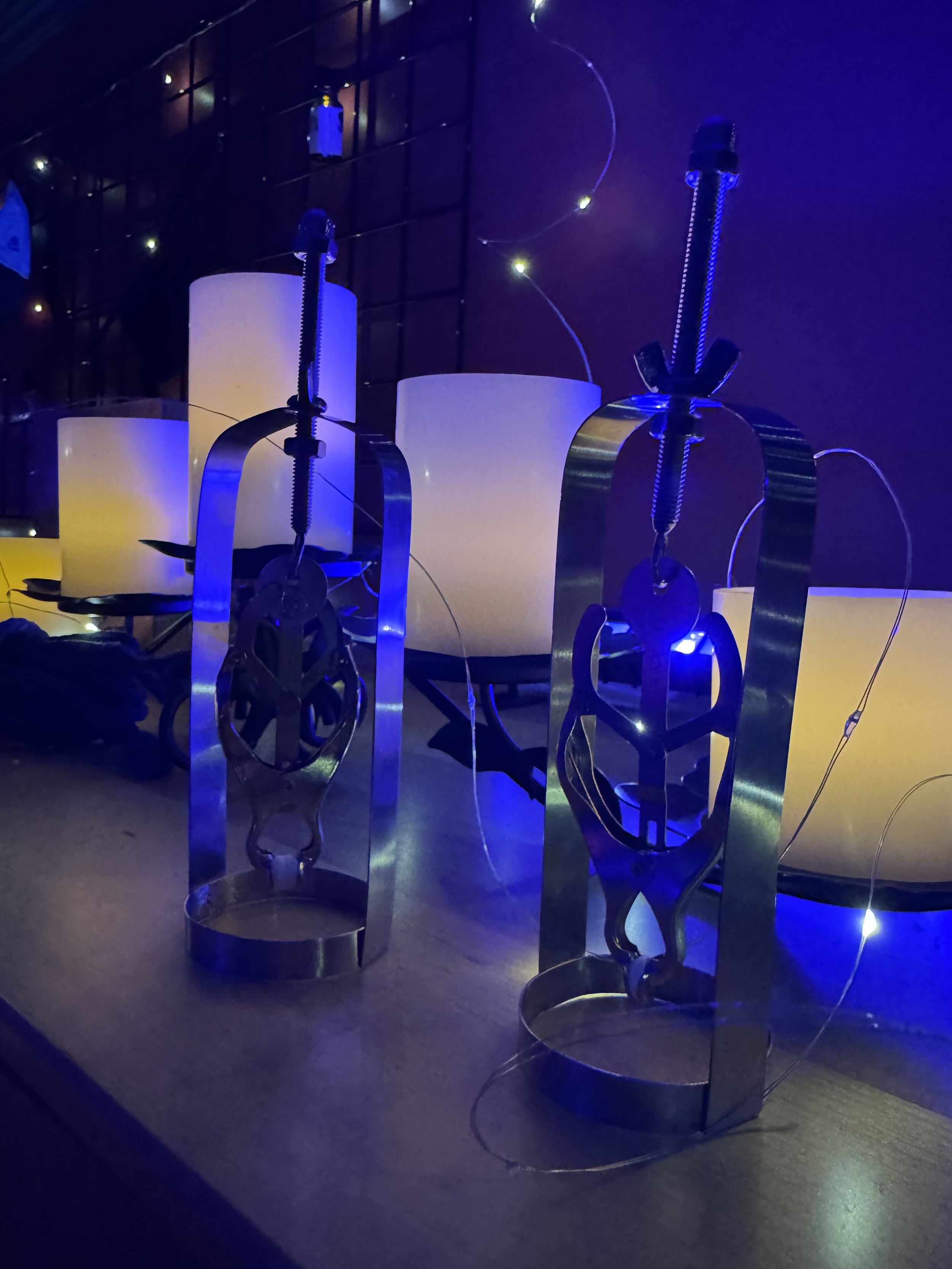 Two metallic bottle openers with a stylized human figure design in front of glowing white lamps on a table, illuminated by blue and purple lighting.