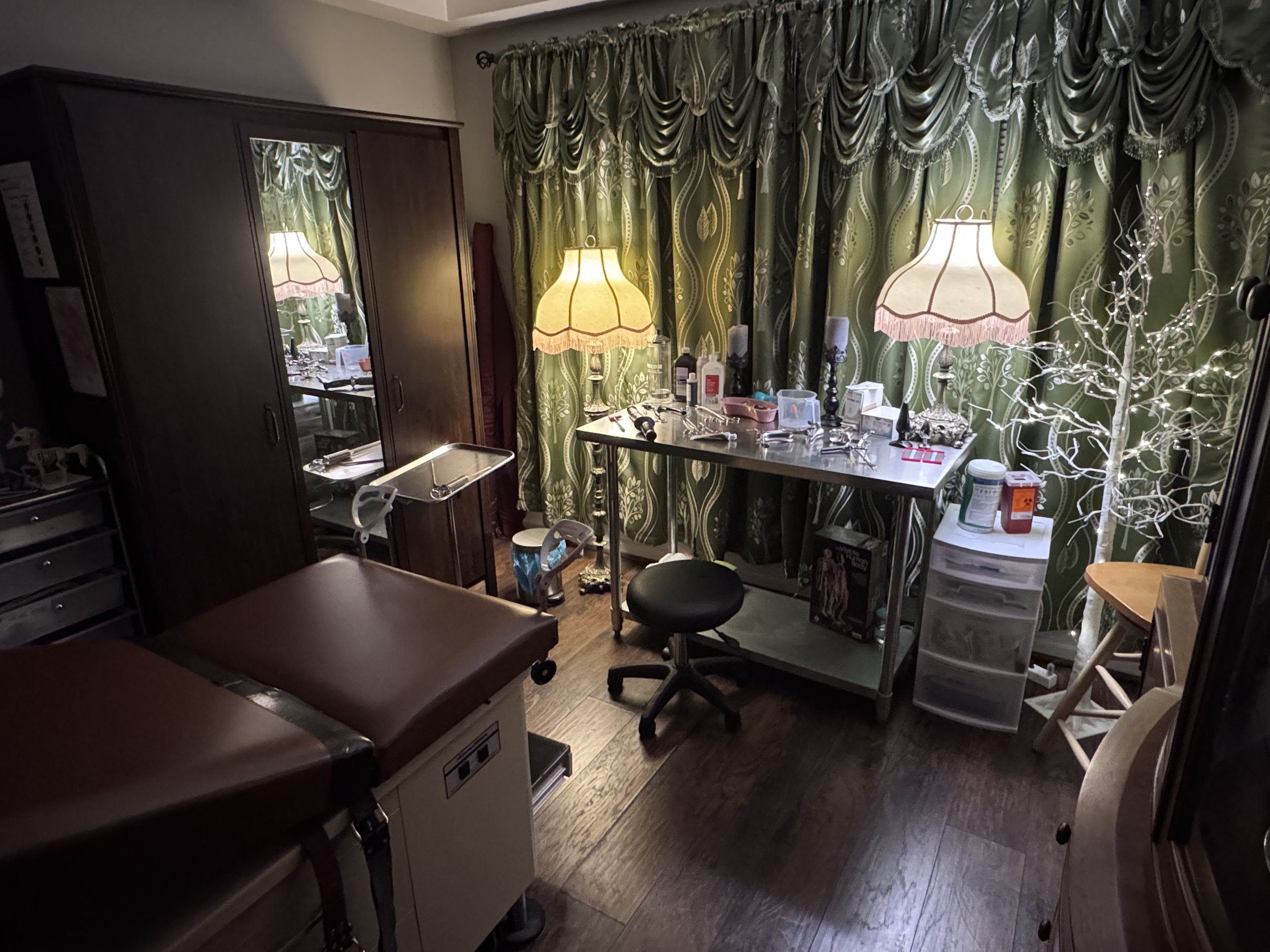 A room with green patterned curtains, two bedside lamps with decorative shades, a white plastic storage drawer, a black stool, and a table cluttered with various items including bottles and a pair of glasses. There is a white tree-shaped decorative l