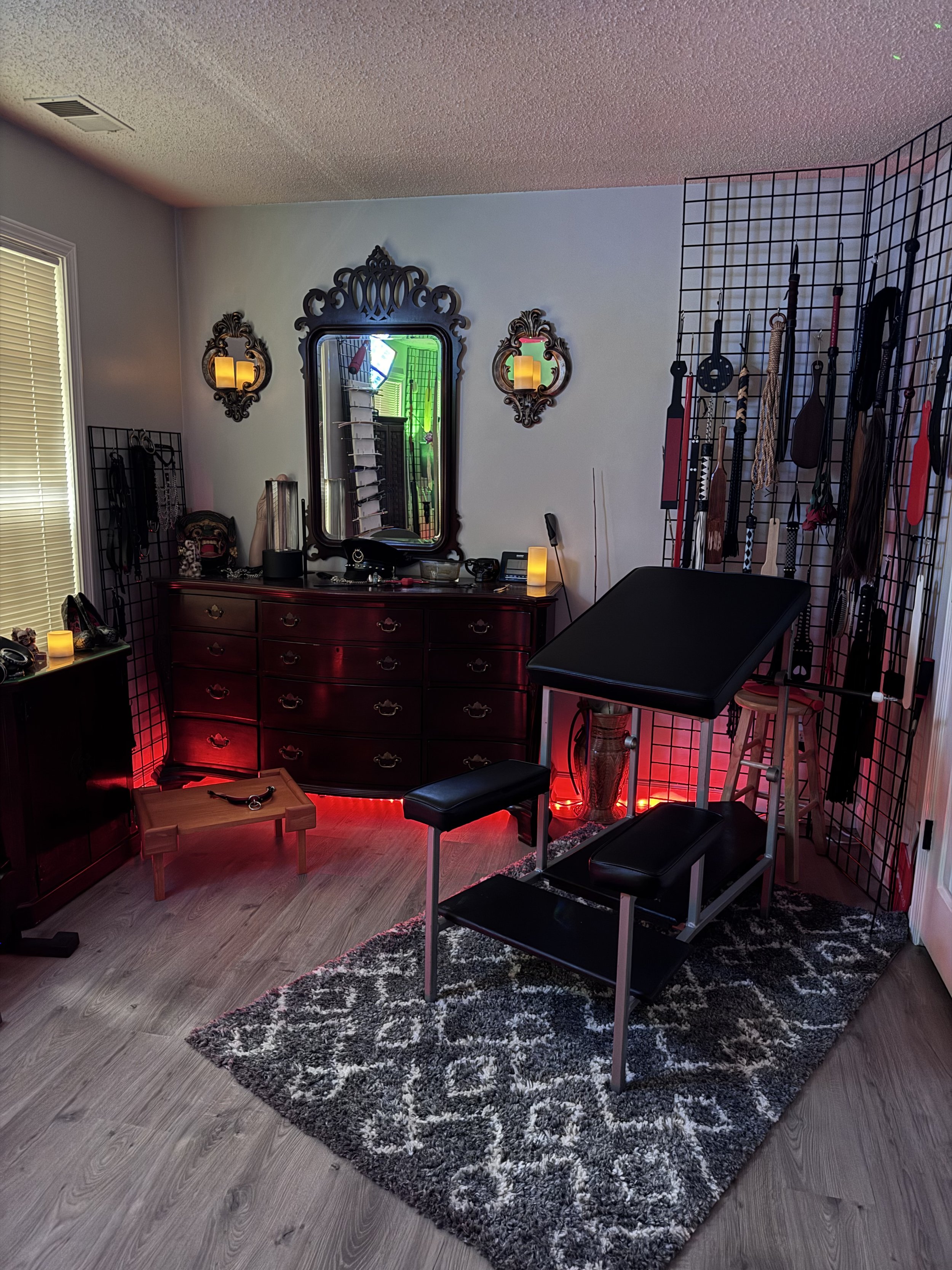A tattoo or medical examination room with a black padded table, a black grid wall display with tools, and a vintage dark wood dresser with ornate mirror and wall sconces. There are neon and LED lights, a rug, and various small items on furniture.