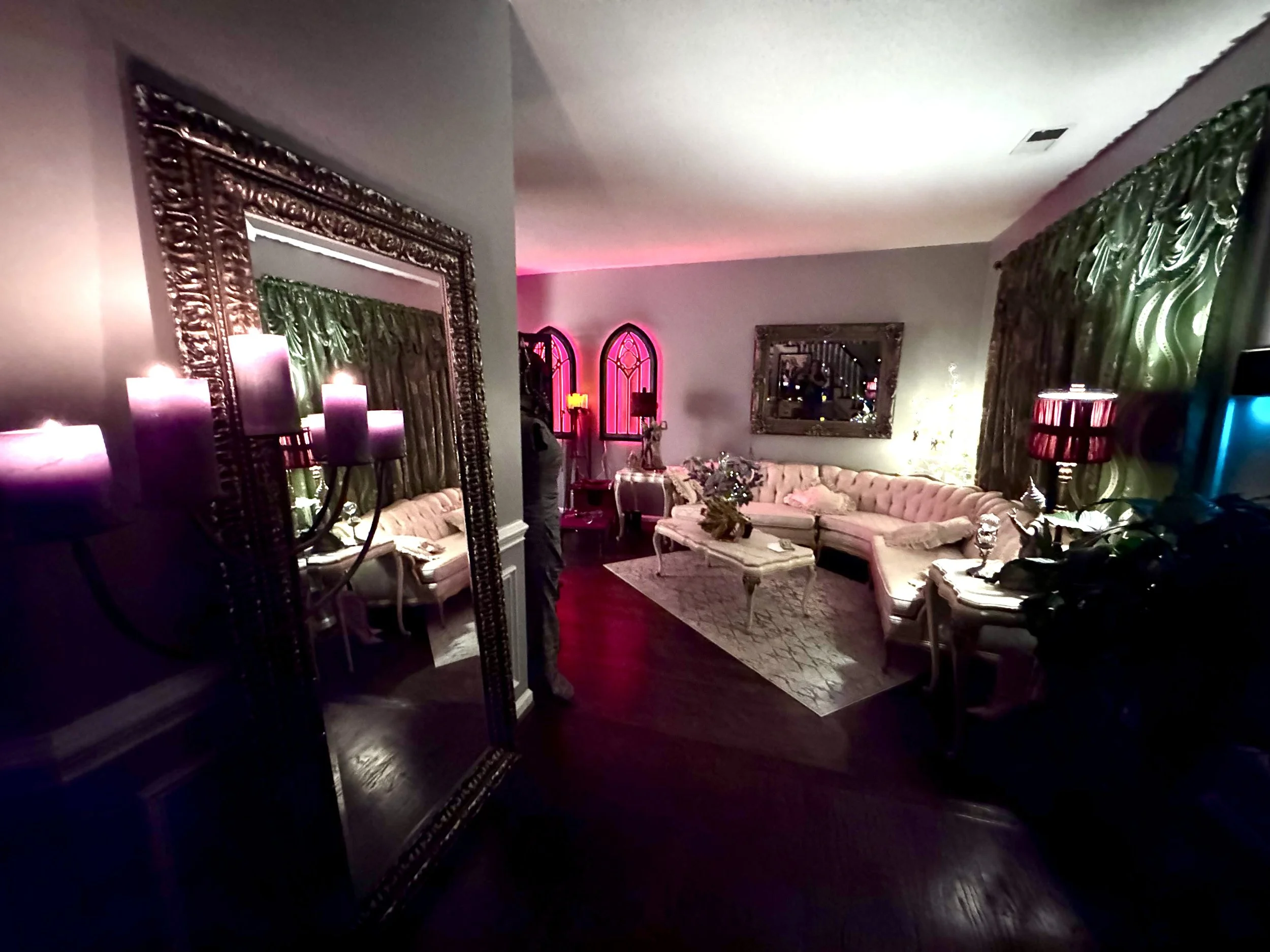 A living room with vintage style furniture, pink sectional sofa, ornate mirror, side tables, lamps, candles, large mirror, and decorative curtains, illuminated with colorful lighting.