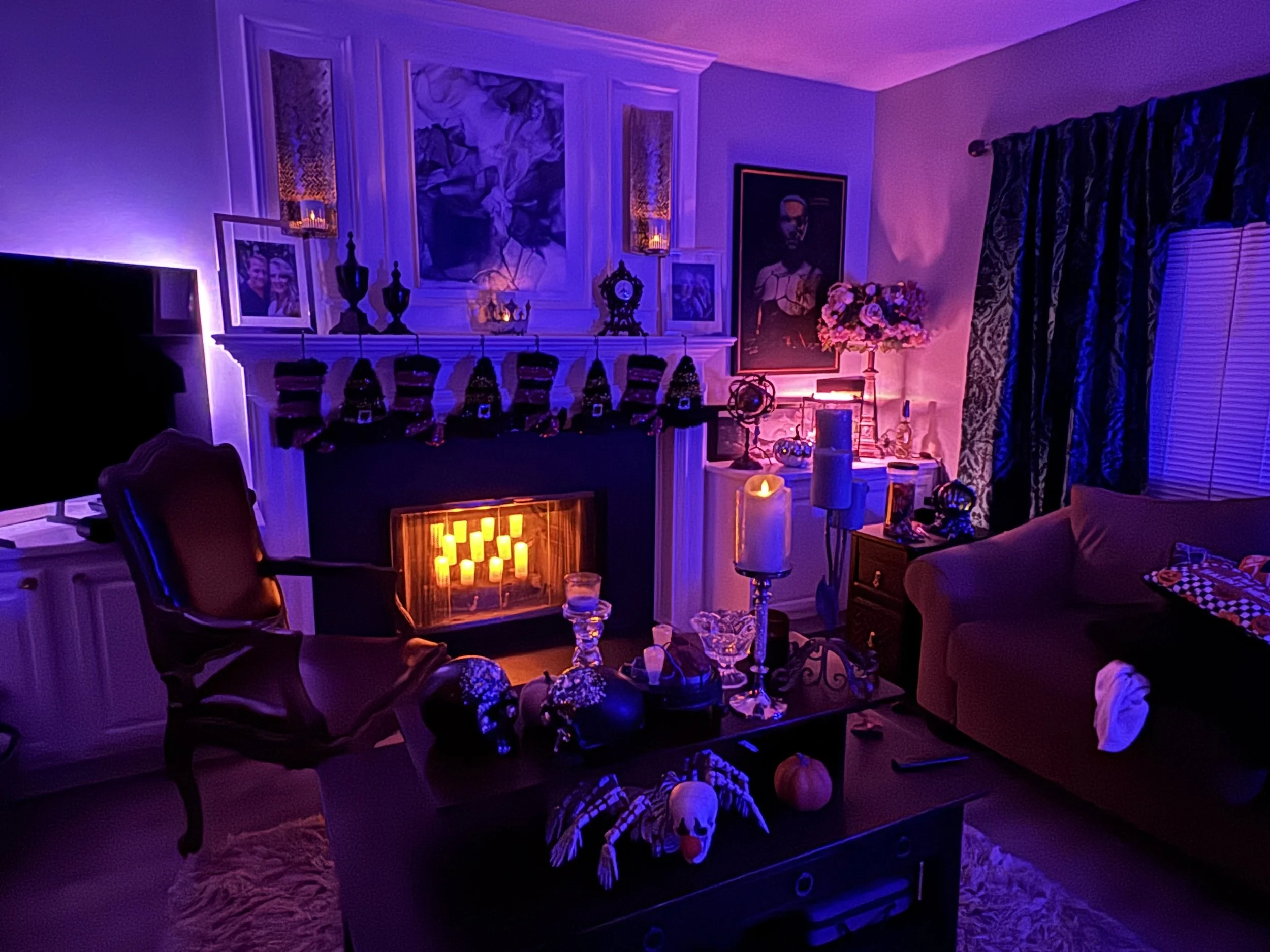 Living room decorated with Halloween and Día de los Muertos items. There is a fireplace with flickering candles, skull decorations, and a rocking chair. The room is illuminated with purple and orange lighting, with picture frames, flower arrangements