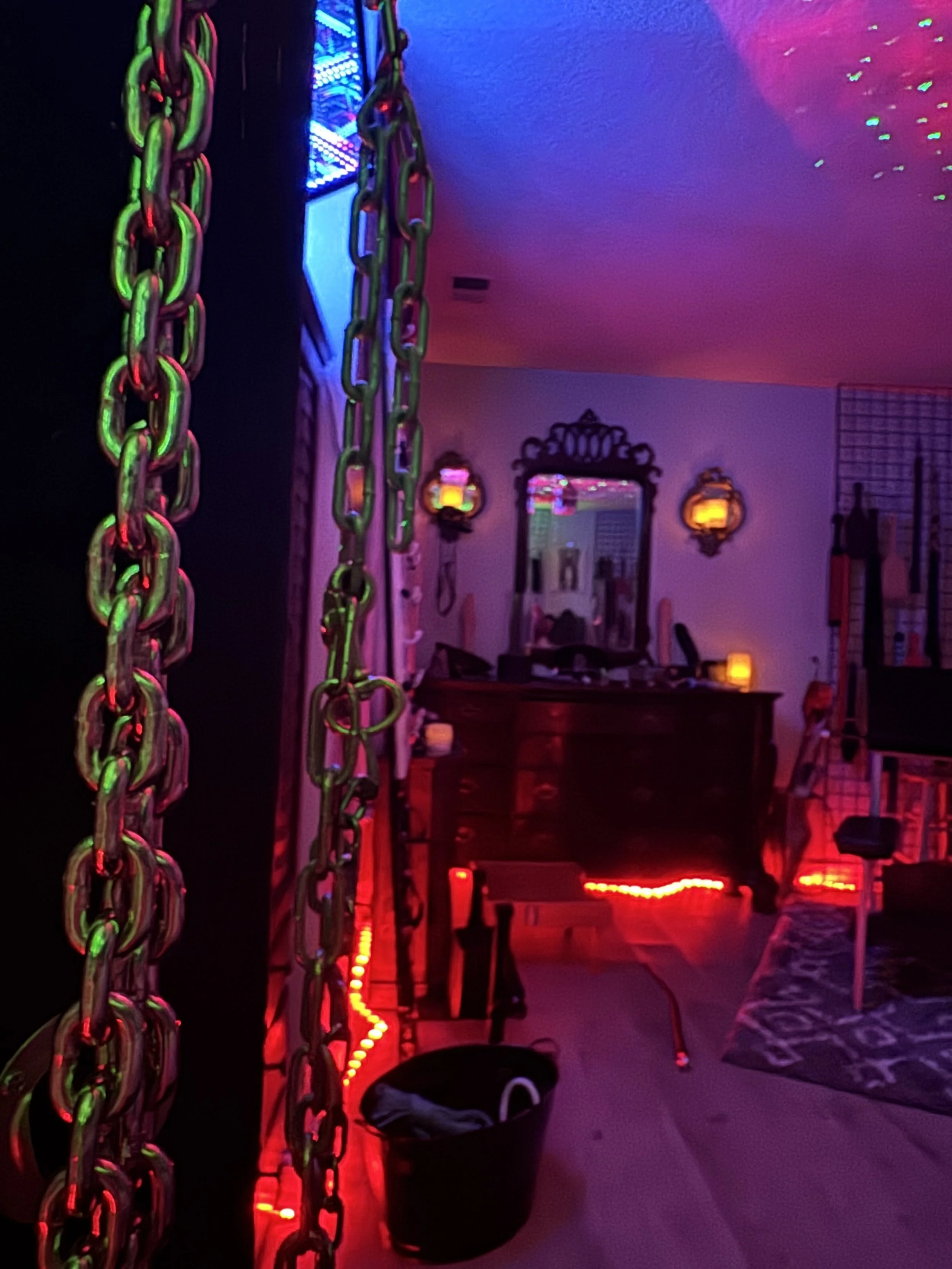 A room decorated with neon and LED lights, including neon chains hanging in the foreground, a mirror, a dresser, and a chair. Purple, pink, and green lighting create a vibrant, party-like ambiance.