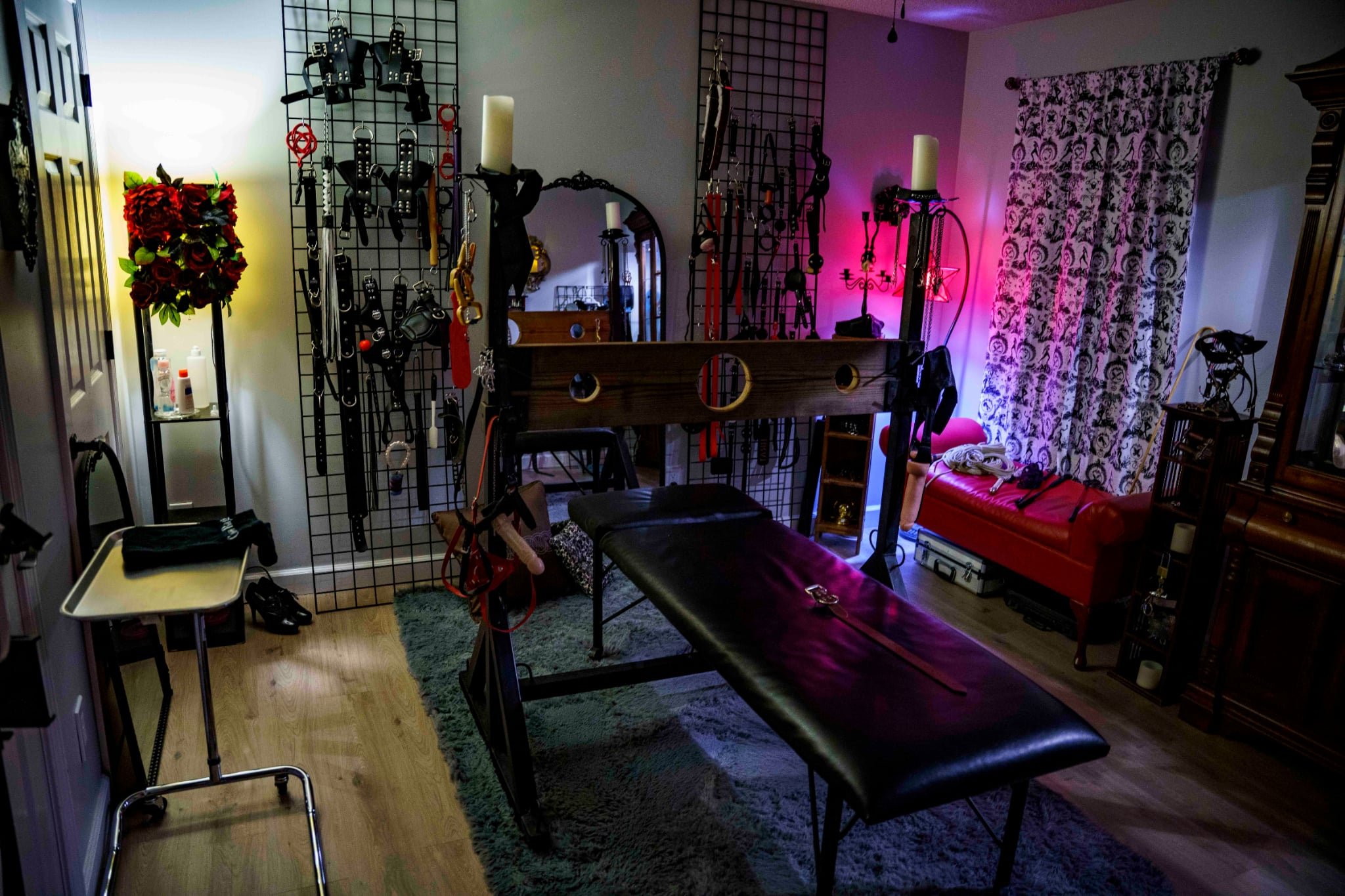 A room with BDSM equipment, including whips, cuffs, and restraints on a hanging grid, a massage table in the center, and a red leather bench with a blanket on it. There are candles, lamps, and decorative curtains, creating an intimate atmosphere.