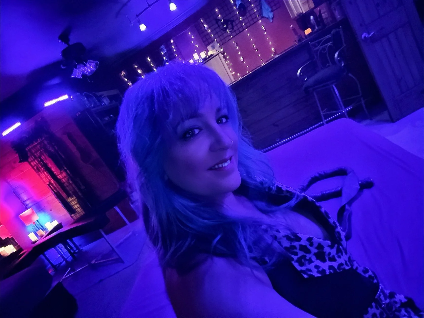 A woman with curly blonde hair smiling at the camera in a dimly lit bar or lounge with blue and purple lighting, decorated with string lights and candles.