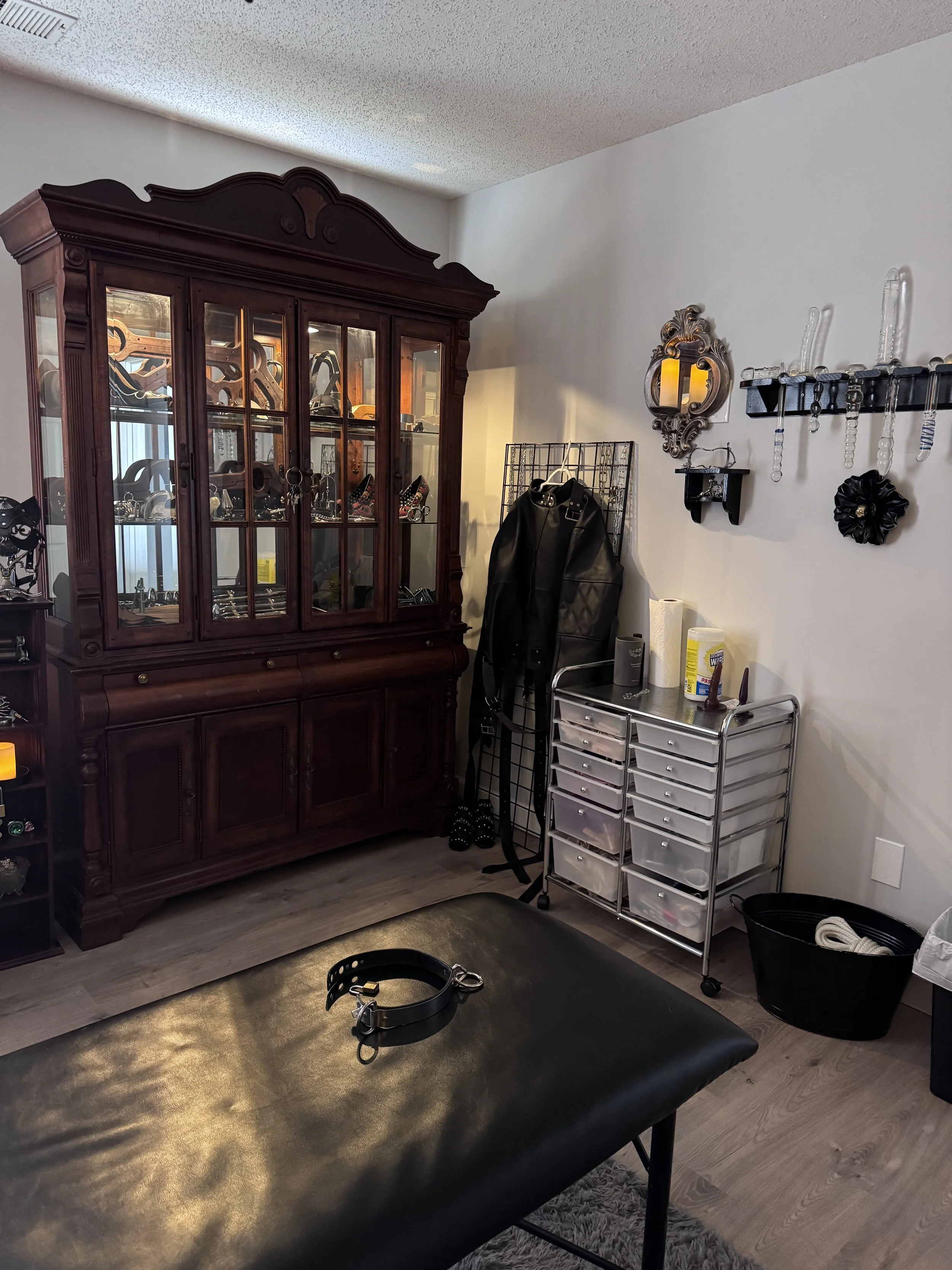 A cozy room with a large wooden display cabinet filled with jewelry and decorative items. There is a black leather bench with a collar on top, a rolling storage cart with supplies, and various accessories on hooks and shelves on the wall.