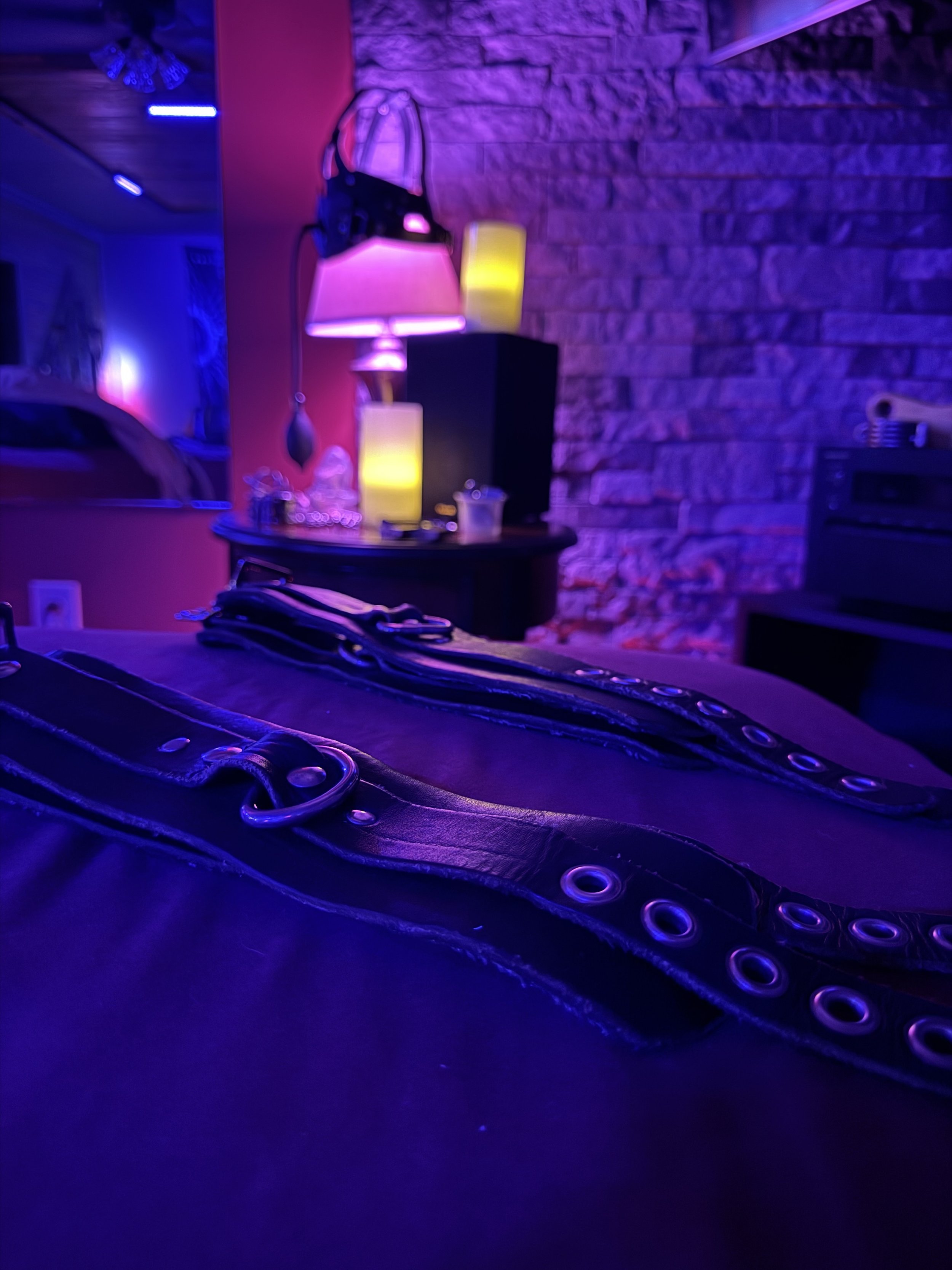 Close-up of leather belt with metal eyelets on a dark surface in a room with colorful neon lighting, including pink and purple hues, with a brick wall and lamps in the background.