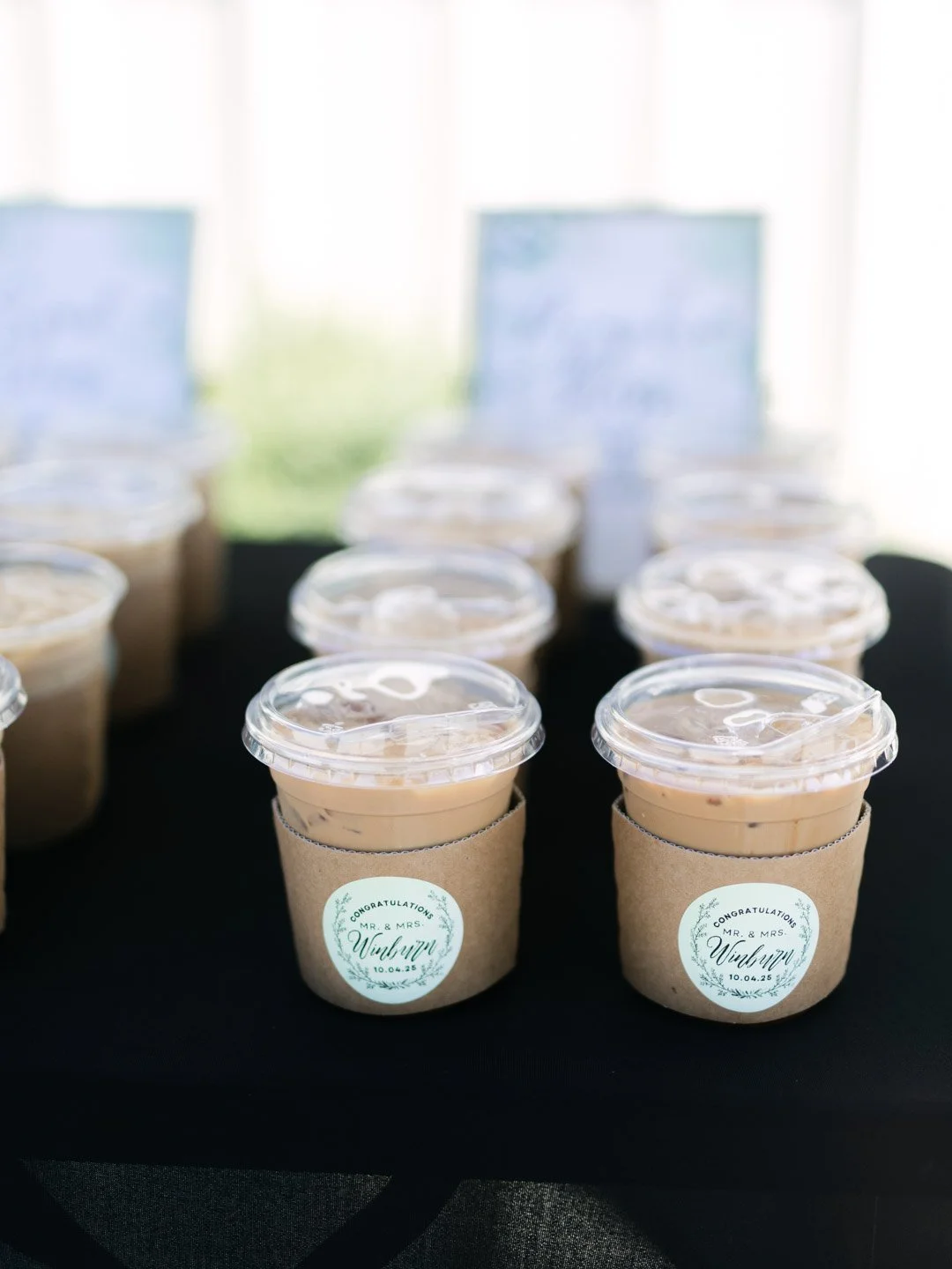 Multiple small cups of creamy dessert with clear plastic lids arranged on a black table, each with a brown paper sleeve that has a wedding-themed label reading "Congratulations Mr. & Mrs. Winbryn 10.04.25."