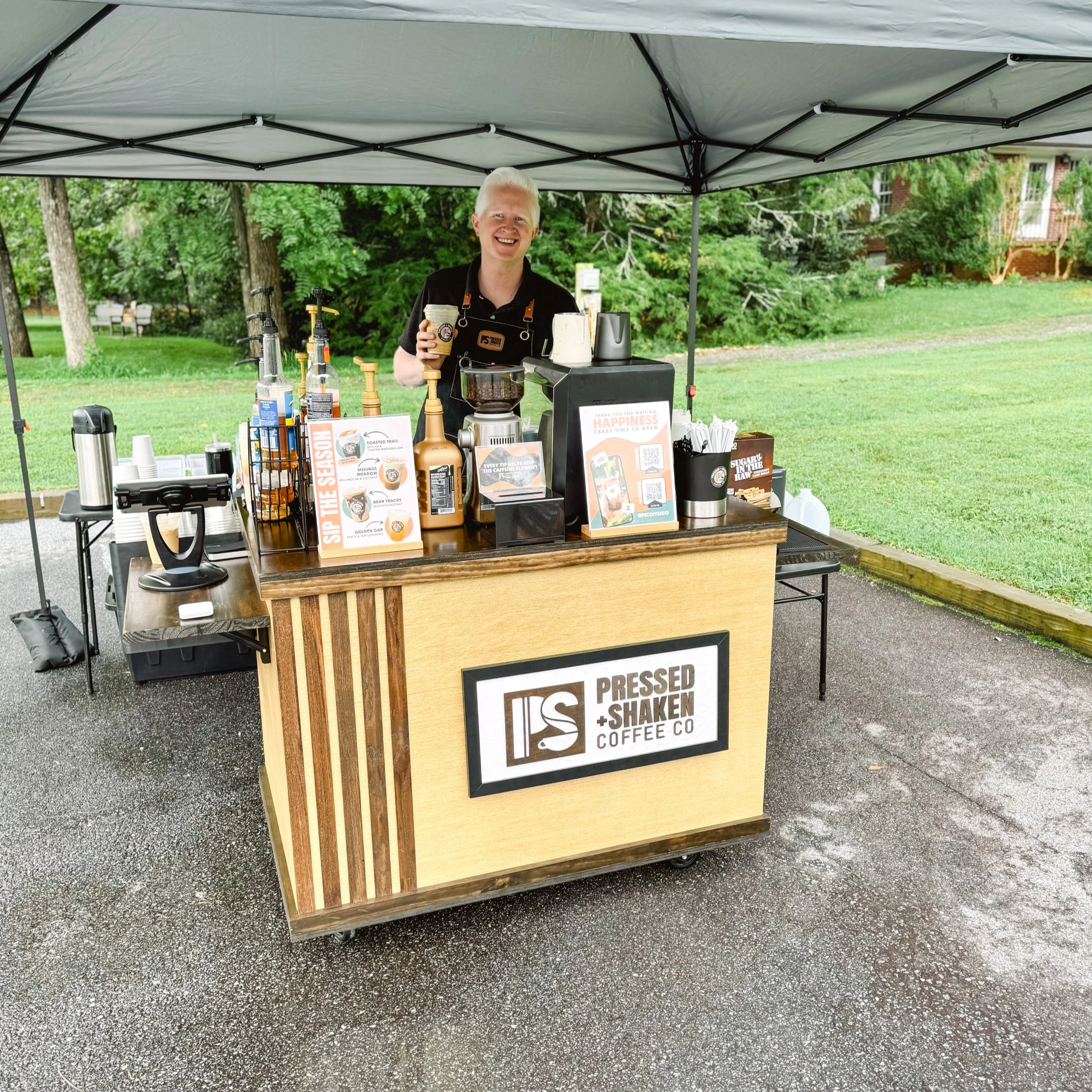 A smiling man standing behind a coffee stand under a canopy, with a green park in the background. The stand has a sign that reads 'Pressed + Shaken Coffee Co' and is equipped with coffee-making supplies and promotional materials.